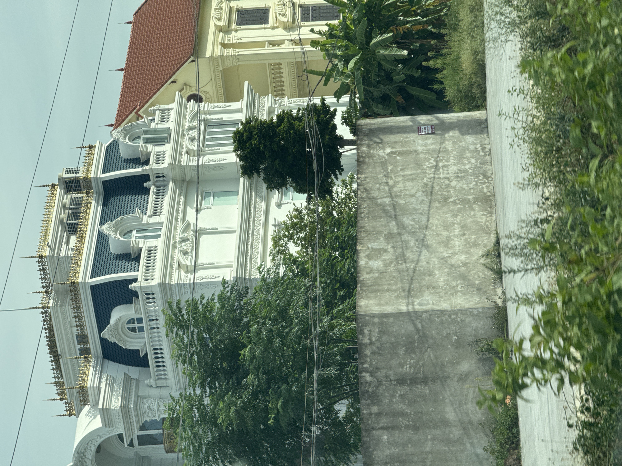 The image depicts a street view of ornate white and yellow buildings in Hoa Lư Ward, Vietnam. The foreground shows a concrete wall and blurred greenery, suggesting the photo was taken from a moving vehicle. The buildings are multi-story structures with intricate architectural details, including mansard roofs with dark blue tiling, decorative white molding, and gold accents. One building is predominantly white, while the adjacent one is yellow with a red tiled roof. Several trees, including palm trees, are visible behind the wall and around the buildings. Overhead, power lines are present. The sky is overcast.