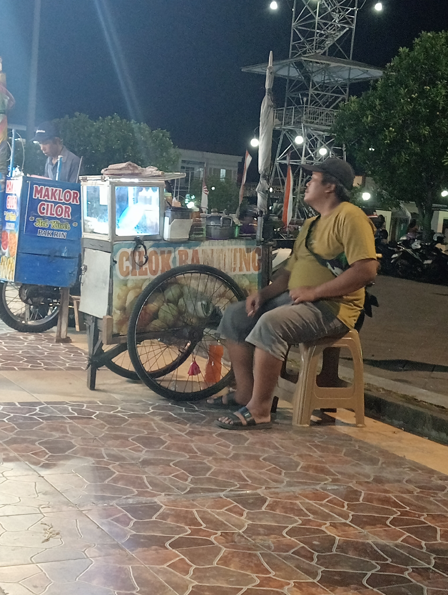 The image depicts an outdoor scene at night in Demak, Indonesia, likely a food stall area. A man wearing a yellow t-shirt and grey shorts sits on a plastic stool, looking to his right. He has a black baseball cap on his head and a small bag slung across his body. Beside him is a food cart with a glass display case showcasing food items. The cart has large wheels and signage in Indonesian that reads "MAKLOR CILOR" and "CIZOK PAK RIN". In the background, a tall metal tower structure with lights is visible, along with trees and other people standing or walking. The ground is paved with hexagonal brown tiles. Motorcycles are parked in the background.