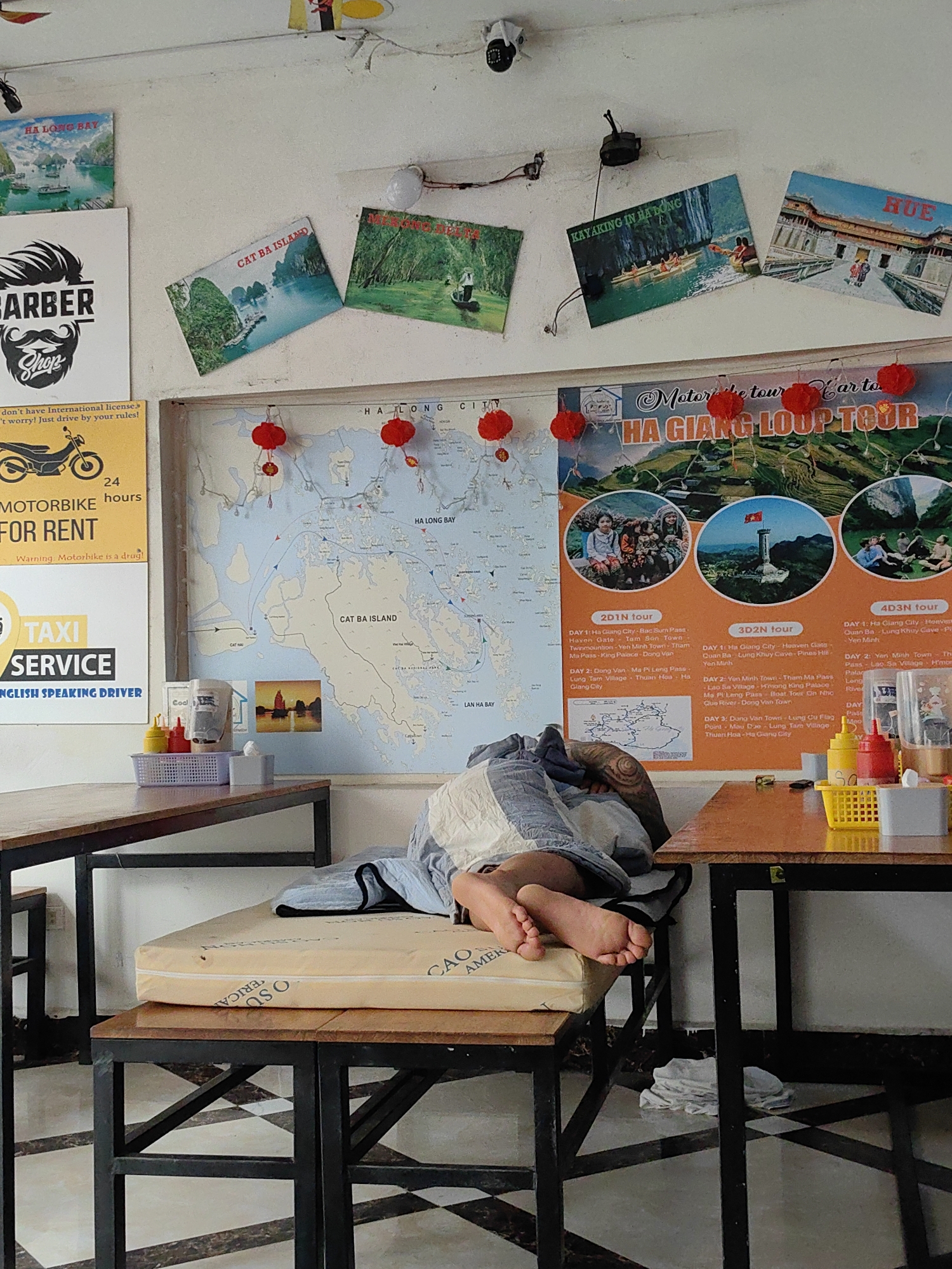 A medium shot captures a scene inside what appears to be a business establishment in Bai Chay Ward, Vietnam. In the foreground, a person is lying on a cushion placed on a wooden table. The individual's bare feet are visible. The table is positioned next to another wooden table with condiments on top. The floor has a black and white checkered pattern. The walls are adorned with maps and tourist posters advertising motorbike tours and local attractions like "Ha Giang Loop Tour" and "Cat Ba Island." A poster advertising a barber shop and motorbike rentals is also visible. The environment suggests a local business catering to tourists in Bai Chay Ward.