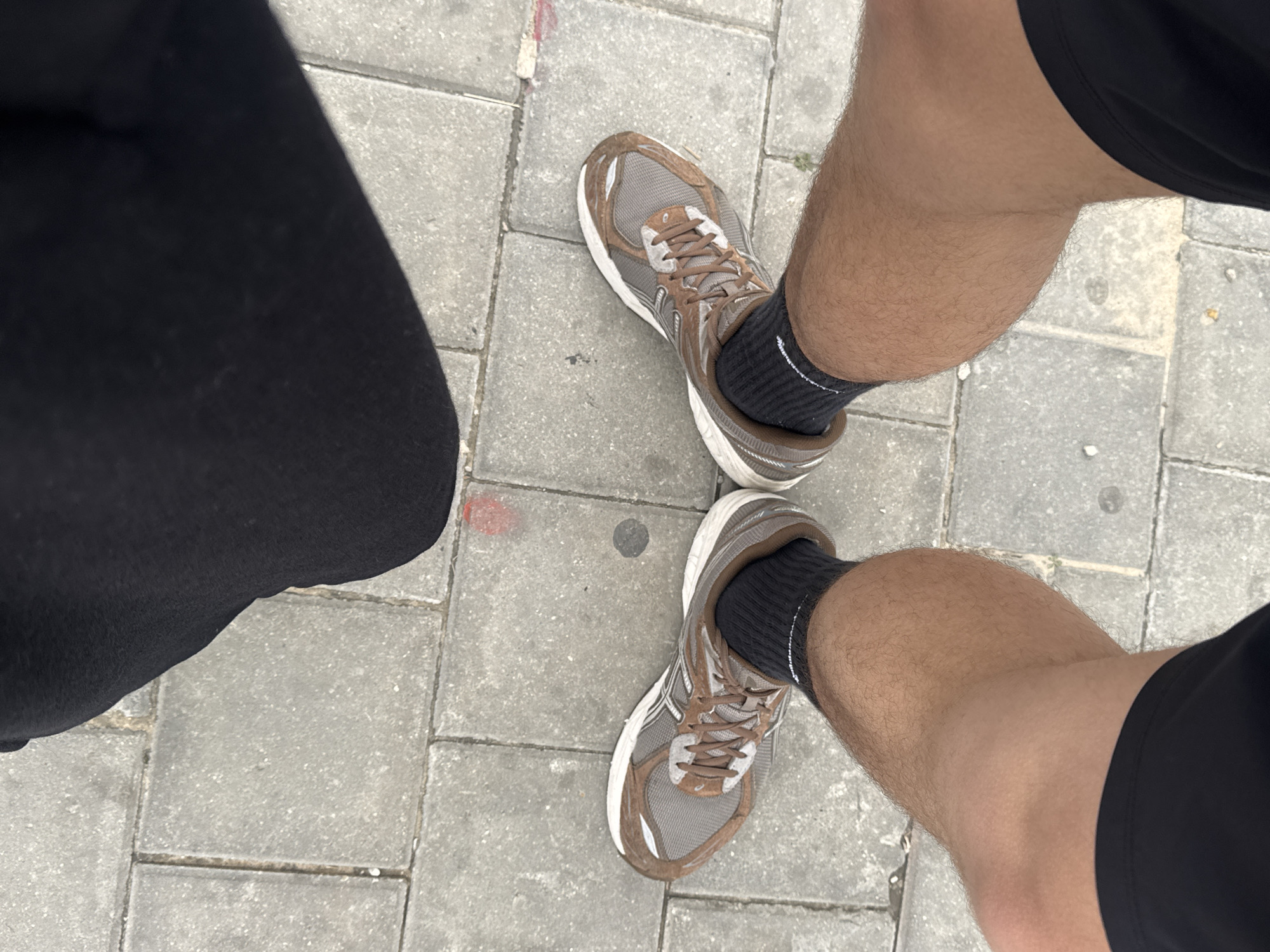 A first-person, downward perspective image displays the lower extremities of an individual standing on a paved outdoor surface in Tel-Aviv, Israel. The subject's two legs, exhibiting moderate hair growth, are visible from approximately mid-thigh. They wear black athletic shorts and black ankle-length socks, each featuring a single white stripe at the cuff. The feet are encased in brown and grey athletic shoes with white outsoles and white lacing systems. The ground consists of grey rectangular concrete pavers arranged in a staggered bond pattern. Distinct markings on the pavement include a faded red circular or irregular mark located front-right of the right shoe, a dark grey circular mark positioned centrally between the shoes, and a partial red mark at the far upper-right. The scene is illuminated by natural light, with a shadow falling from the top of the frame, obscuring part of the pavement and the left shoe. The precise location within Tel-Aviv is not discernible from the visual data, beyond being an urban paved area.
