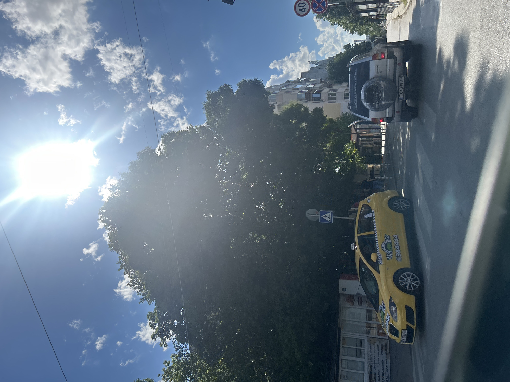 The image captures a sunny day in Varna, Bulgaria, likely in a residential or mixed-use area. The foreground shows a street with a pedestrian crossing. A yellow taxi, identified by the license plate "B 8760 TH" and the phone number "344444" displayed on its side, is stopped at the crossing. To the right of the taxi, a silver SUV with the license plate "TX 4899 AP" is also visible. Beyond the vehicles, a large, leafy tree dominates the left side of the frame, casting shadows on the street. In the background, a multi-story building with balconies stands on the right, and a street sign indicating a 40 km/h speed limit is present. The sky is bright blue with scattered white clouds, and the sun is high and shining intensely, creating lens flare and washing out parts of the image. There are power lines visible across the top of the frame. A few indistinct figures are present on the sidewalk in the mid-ground. The overall scene suggests a typical urban street environment under clear, sunny weather.
