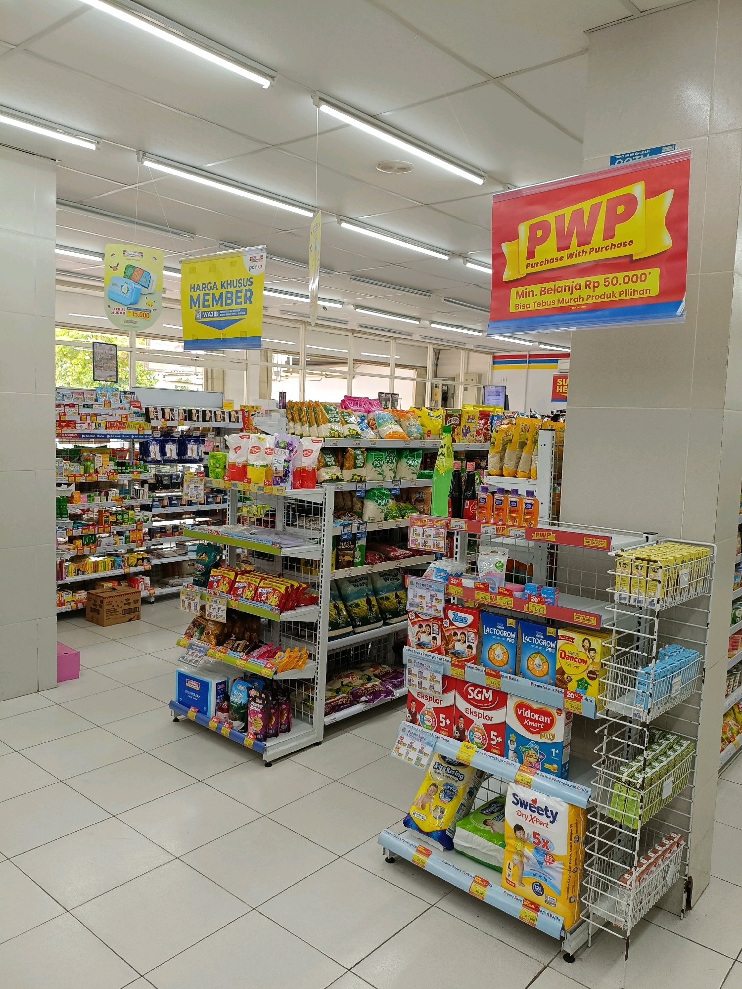 The image depicts the interior of a minimarket, likely in Maguwoharjo, Indonesia, judging by the signage. Fluorescent lights illuminate the brightly lit space, reflecting off the white tiled floor and the rows of shelves stocked with various consumer goods. The shelves are filled with packaged food items, beverages, and household products. Promotional banners are hung from the ceiling and attached to the shelves, advertising special offers like "HARGA KHUSUS MEMBER" (Special Price for Members) and "PWP Purchase With Purchase" with minimum spending requirements. The items on display include boxes of milk powder, snacks, bottled drinks, and other everyday essentials. There are no people visible in the frame. The overall impression is one of a well-organized retail environment.