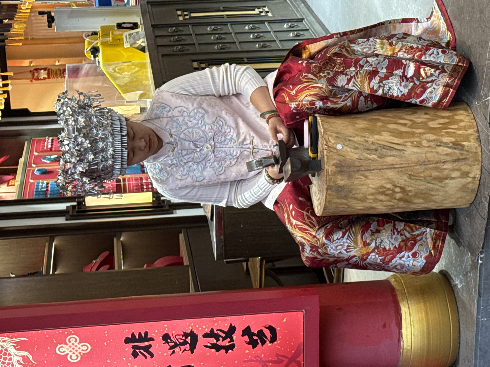 A woman wearing a silver headdress and a light pink embroidered shirt sits on a red patterned cloth draped over a large wooden cylinder. She is working with a hammer and a small anvil. The scene takes place indoors, likely a store, as evidenced by display shelves in the background. A cabinet with drawers sits behind her. A sign with Chinese characters is visible in the foreground, and a portion of what seems to be a red and gold pillar is also in view. The exact location is Shapingba District, China.