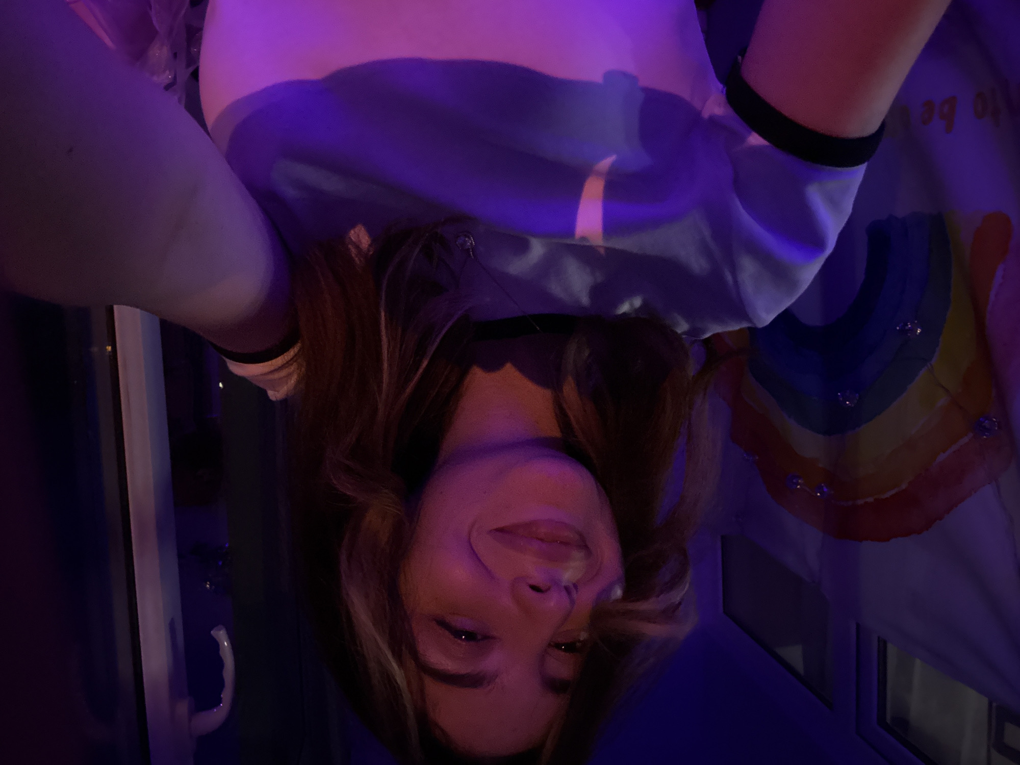 A female with shoulder-length brown hair is pictured in an indoor setting. She is wearing a white shirt with a blue design and black wristbands. A rainbow-colored object is partially visible in the upper right corner. The scene is lit with blue and purple lighting. The location is unknown.