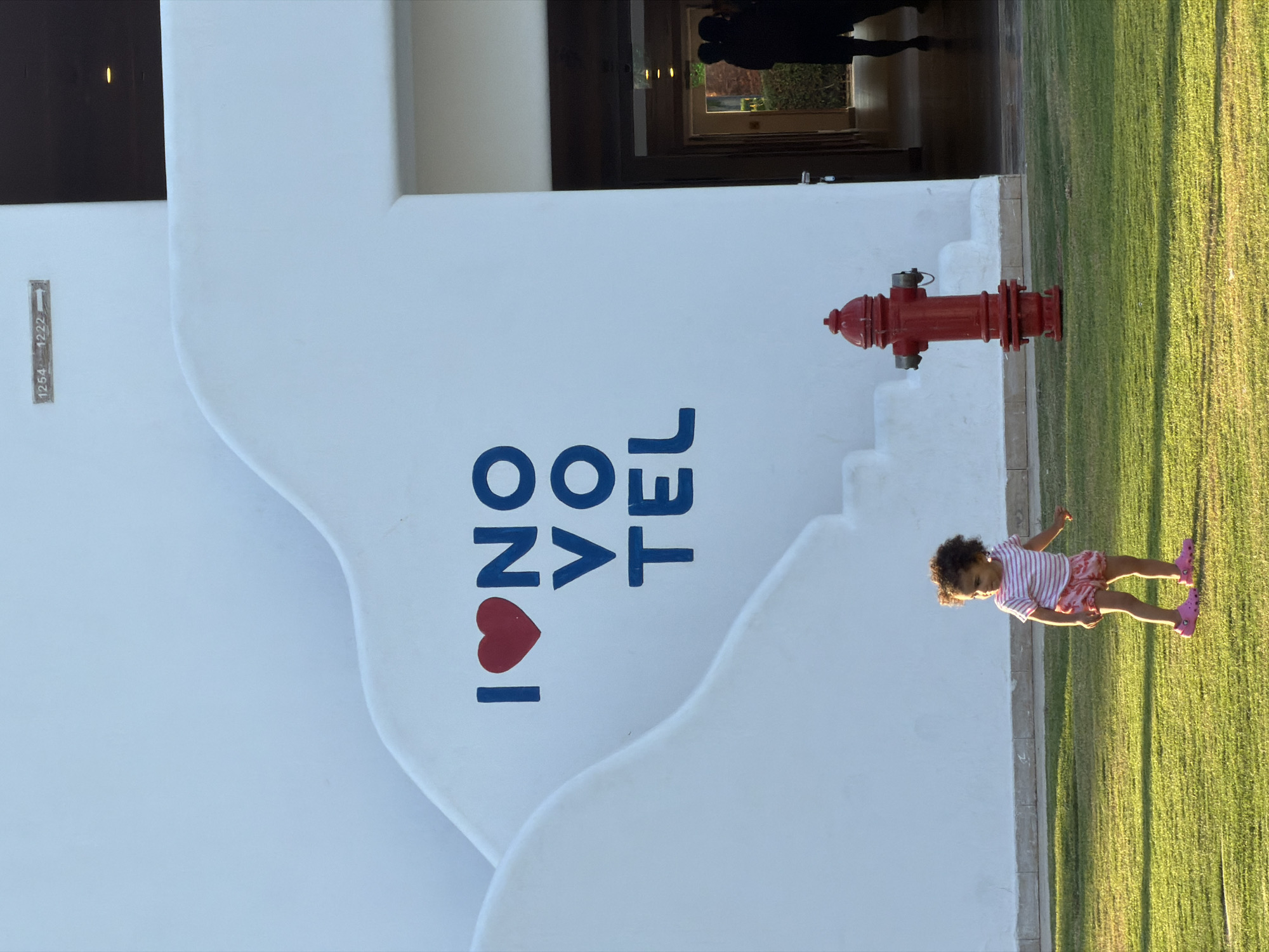 The image shows a low-angle, full-shot photograph of a young child standing on a manicured lawn in front of a white stucco building. The child, appearing to be a toddler, is wearing a pink and white striped t-shirt and patterned pink shorts. They are looking to their right, with their left arm extended slightly. The building has a distinctive curved architectural style and features the words "I ♥ NO VO TEL" painted in blue and red on its facade. To the right of the text, a bright red fire hydrant stands on the lawn. In the background, an open doorway leads into a dimly lit interior, where the silhouettes of two individuals are visible. A sign with numbers "1254 - 1222" and an arrow is mounted high on the building's wall. The scene appears to be set in a resort or hotel complex. Based on the architectural style and the "NOVOTEL" branding, this location is likely the Novotel Sharm El Sheikh, located in Sharm El Sheikh, Egypt.