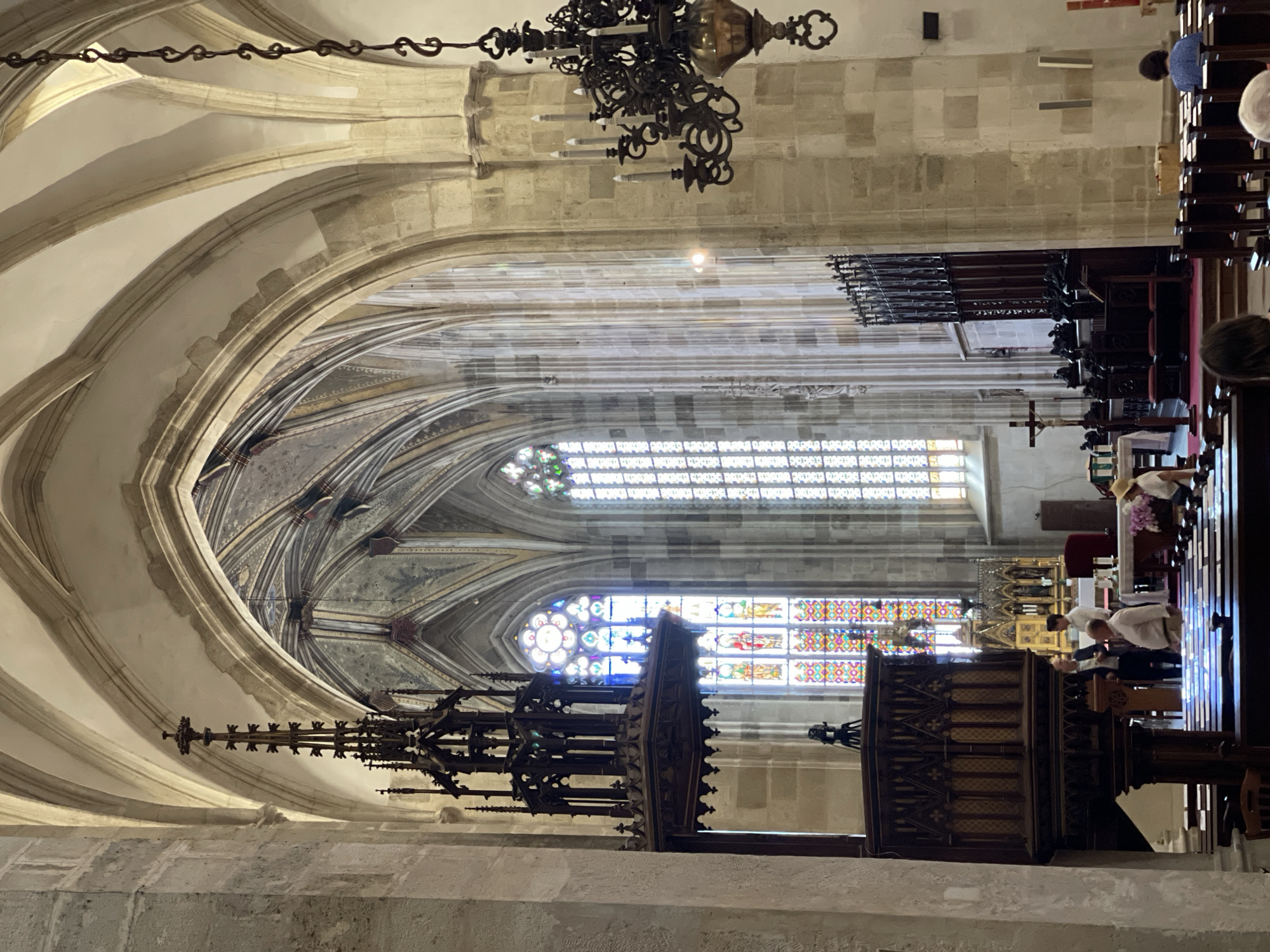 The image depicts the interior of St. Martin's Cathedral in Bratislava, Slovakia. The high-vaulted ceilings and stained-glass windows are prominent architectural features. In the foreground, a large wooden pulpit is visible on the left. Several individuals are present, some standing near the pulpit and others seated in the pews. A chandelier hangs from the ceiling on the right side of the image. The cathedral's interior is characterized by stone columns, ornate woodwork, and religious iconography, including a crucifix and an altar area visible in the background. The lighting suggests it is daytime, with light filtering through the stained-glass windows and illuminating the interior.