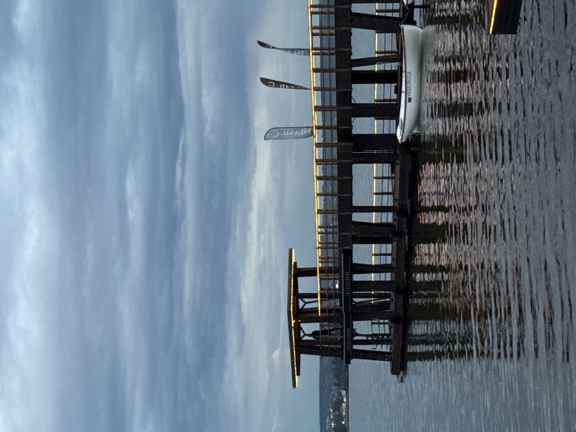 The image depicts a wooden pier extending over a body of water, possibly a lake or river, in Votkinsk, Russia. The sky is overcast with a mixture of light and dark grey clouds. The pier is illuminated by yellow lights along its edges and on a small elevated structure at one end. A silhouette of a person is visible standing on this elevated section of the pier. Several tall, narrow flags are attached to the railings of the pier, some displaying writing. A small white rowboat is moored to the pier, with visible writing on its side. The water reflects the lights from the pier, creating shimmering patterns. In the background, a distant shoreline with buildings and trees is faintly visible. The overall scene suggests an evening or dusk setting.