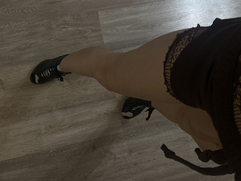 A high-angle shot shows legs and feet on a wood-look floor. The subject wears dark athletic shoes and a black garment with a ripped or netted edge, possibly a skirt or shorts. The legs are fair-skinned, slightly angled, with one foot pointing away and the other toward the camera. The focus is somewhat soft, giving the image a slightly dreamy quality. The lighting is muted and cool.