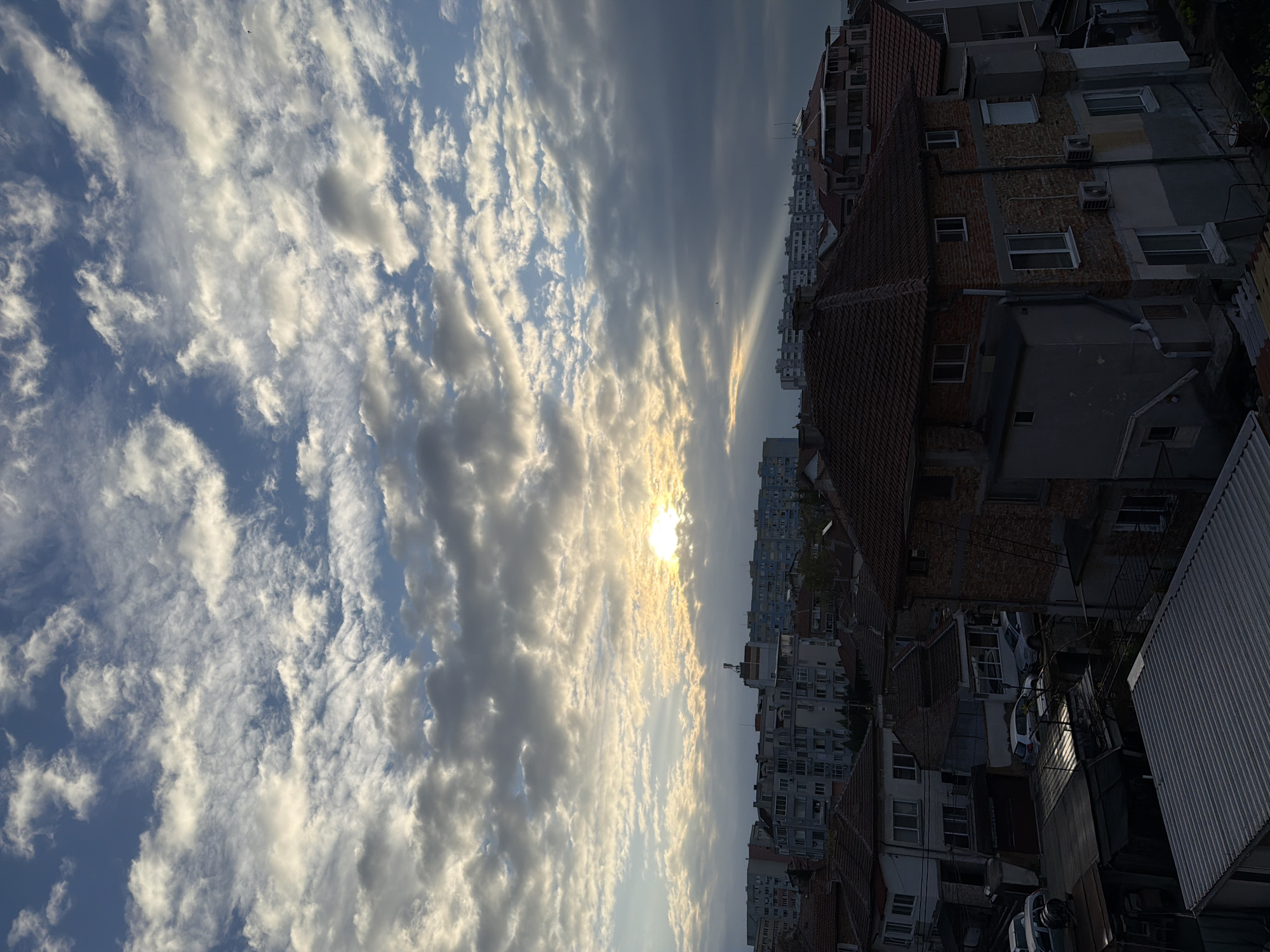 Here is a forensic description of the image:
The image captures a cityscape under a sky filled with altocumulus clouds, some obscuring the sun. The clouds are predominantly white and gray, with patches of blue sky visible. Sunlight is diffused through the clouds, creating a bright glow in the central area of the sky.
On the right side of the frame, there are buildings with brick and concrete facades. Some of these buildings have visible windows, air conditioning units, and tiled roofs. Further in the background, taller buildings are seen, partially obscured by the closer buildings.
In the foreground, there are rooftops and other building structures. Cars are visible parked along the street.
The location in the city is unknown. The orientation of the image is rotated ninety degrees.
