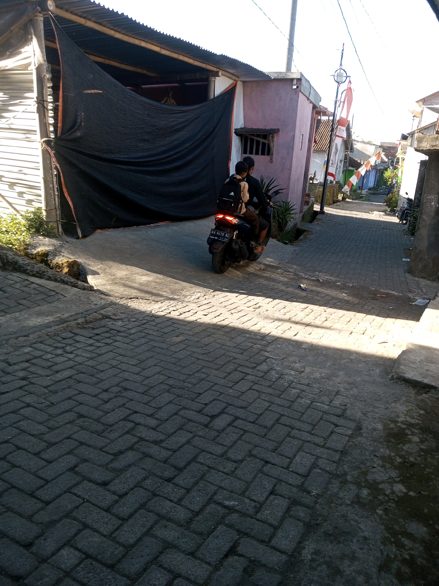 A motorcycle with two occupants is parked on a brick-paved alleyway in Andongsili, Indonesia. The driver, wearing dark clothing and a backpack, is seated. The passenger, also in dark clothing and wearing a backpack, is behind the driver. The motorcycle has a license plate that reads "AA 6479 OZ". The alleyway is lined with buildings, one of which is partially covered by a large black tarp. Indonesian flags are visible on poles in the background, suggesting a festive occasion. The sun is casting strong shadows across the alley.