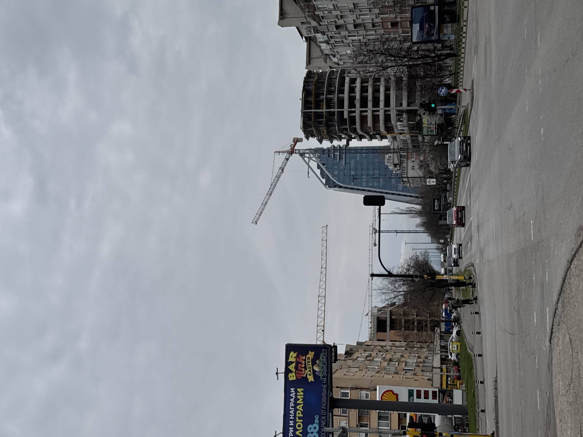 The image captures a street scene in Sofia, Bulgaria, on a cloudy day. The street is lined with buildings of varying heights and styles, including some under construction. Several construction cranes are visible in the skyline. There is a street intersection with traffic lights. A few vehicles are visible on the road. A pedestrian is standing at the intersection. A billboard is visible in the foreground, advertising a "BAR Link EASTER" promotion. The image presents an urban environment with active construction and everyday street activity.