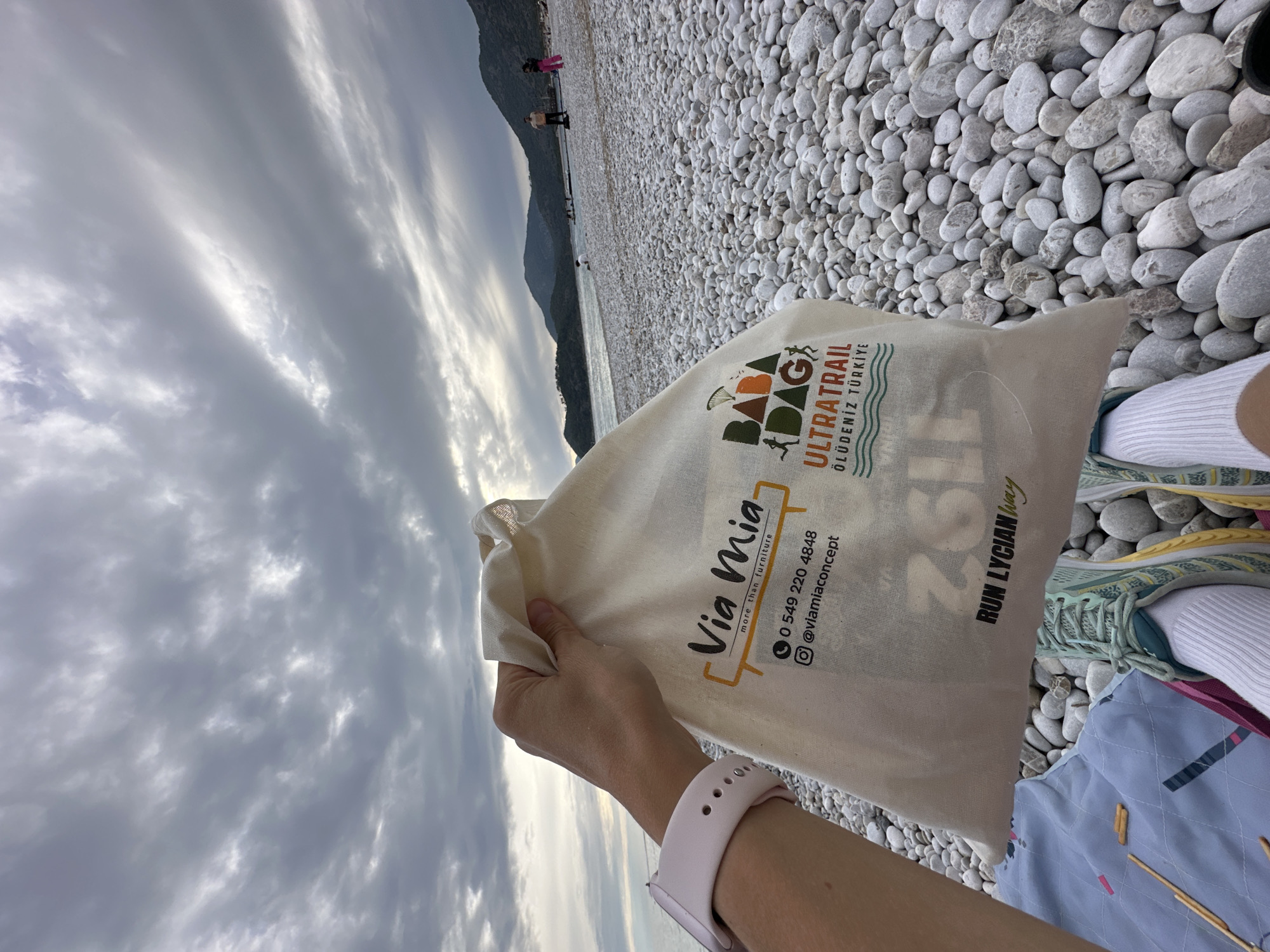 The image shows a first-person perspective of a scene on a pebble beach, likely in Fethiye, Turkey. In the foreground, a person's hand holds a cream-colored canvas tote bag. The wrist wears a light pink smartwatch. Visible below the bag are parts of legs, running shoes with light blue and yellow details, and white socks, suggesting an active individual. The person appears to be resting on a patterned blue and pink mat, with a few small, stick-like snacks (possibly pretzels or breadsticks) scattered nearby.
The tote bag features prominent text and logos. One side advertises "Via Mia more than furniture" along with a phone number (0 549 220 4848) and an Instagram handle (@viamiaconcept). The other side clearly displays event details: "BABA DAĞ ULTRATRAIL ÖLÜDENİZ TÜRKİYE," accompanied by a logo featuring a runner and a paraglider. Below this, "RUN LYCIAN way" is visible, along with a partial number "202," strongly indicating participation or involvement in an ultratrail running event on the famous Lycian Way.
The background reveals an expansive pebble beach leading to a body of water, likely the sea, which stretches towards distant, dark mountains. The sky above is largely overcast with elongated, stratiform clouds, creating diffuse lighting conditions. Several small figures can be seen walking along the beach in the distance. The overall scene captures a moment of rest at a scenic location during or after an ultratrail event in the Ölüdeniz region of Fethiye, Turkey.