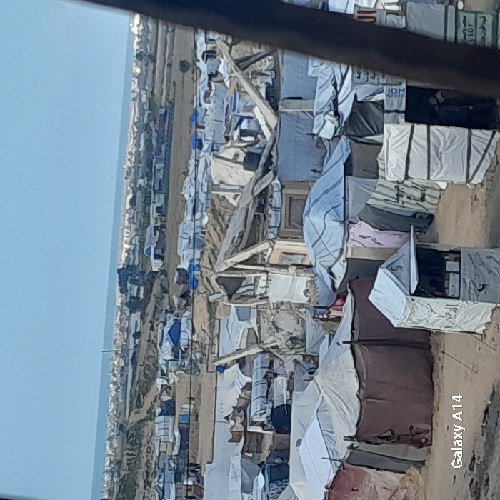 The image captures a sprawling, dense encampment in Khan Yunis, Palestinian Territory, under a clear, light-blue sky. The scene is dominated by numerous makeshift shelters and tents constructed from a variety of materials including white and colored tarpaulins, fabrics, wooden frames, and some corrugated metal. Many structures exhibit signs of rudimentary construction, damage, or partial collapse, with exposed support beams and torn coverings. The ground throughout the encampment appears to be sandy dirt. In the immediate foreground, a small, white, tent-like kiosk with an open flap displaying items is visible, featuring Arabic script on its side. To the right, a partially visible individual stands near another white, box-like structure. The encampment extends extensively into the midground. The distant background reveals a flat expanse leading to what appears to be a coastline or large body of water, beyond which a low-rise urban landscape is discernible. The overall scene depicts a crowded, temporary or semi-permanent settlement.