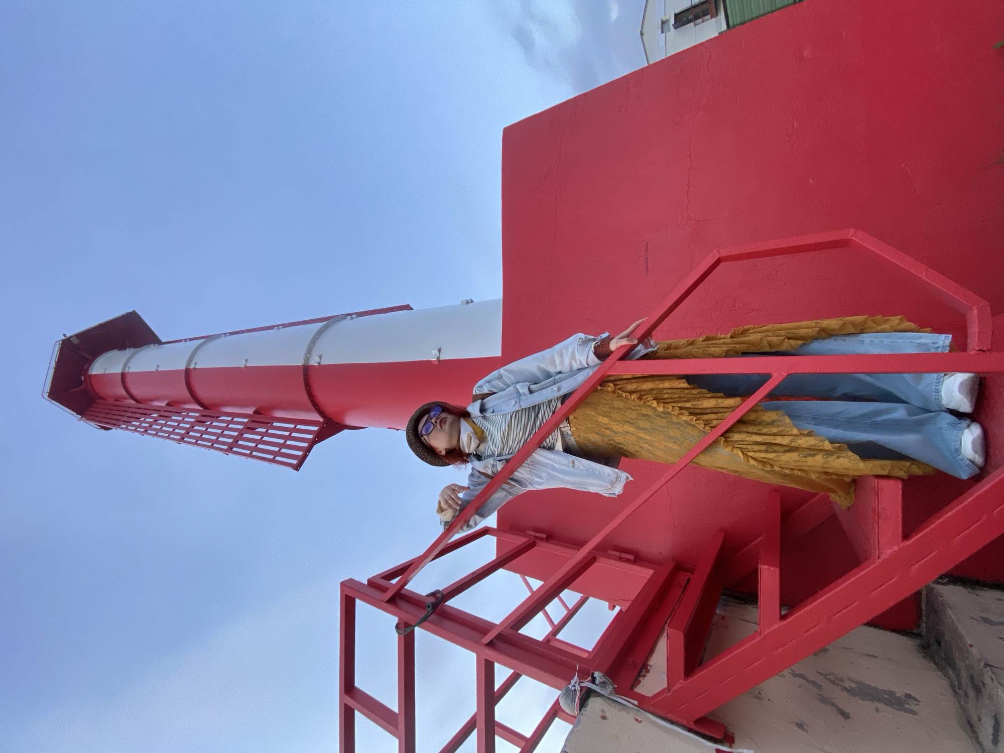 A low-angle shot captures a person lying on red metal stairs attached to a red and white lighthouse-like structure against a cloudy blue sky. The individual wears a hat, sunglasses, a striped shirt under a light denim jacket, blue jeans, and white shoes. A yellow, ruffled garment is also draped over them. The location is likely in Sochi, Russia, based on the distinctive style of the lighthouse and other subtle contextual clues.