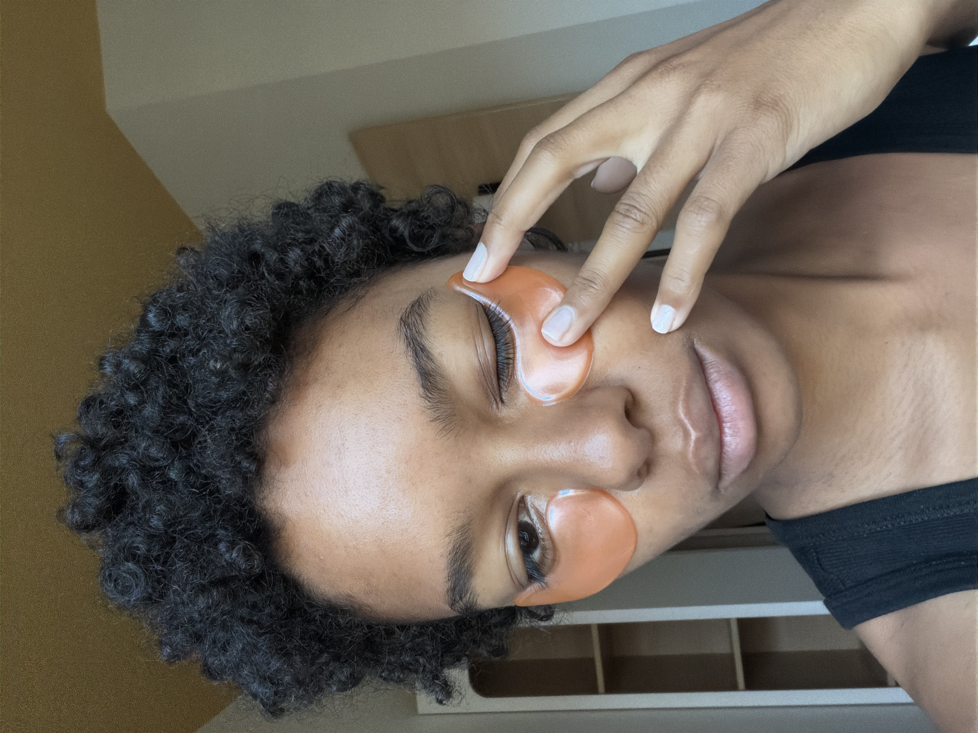 A close-up, head-and-shoulders shot shows a young Black woman with short, dark, curly hair and light skin. She is looking slightly to her left with her right eye open and her left eye partially closed, as if winking. She has two orange under-eye patches on her face. Her left hand is raised to her face, with her index and middle fingers touching the under-eye patch on the left side of her face. She is wearing a black tank top. The background is a plain, yellowish wall with a corner of a room visible on the right, possibly a closet or doorway. A white bookshelf is partially visible in the background to the left. The lighting suggests an indoor setting, possibly a bedroom or bathroom. The image does not contain any information about the city of Bremen, Germany.