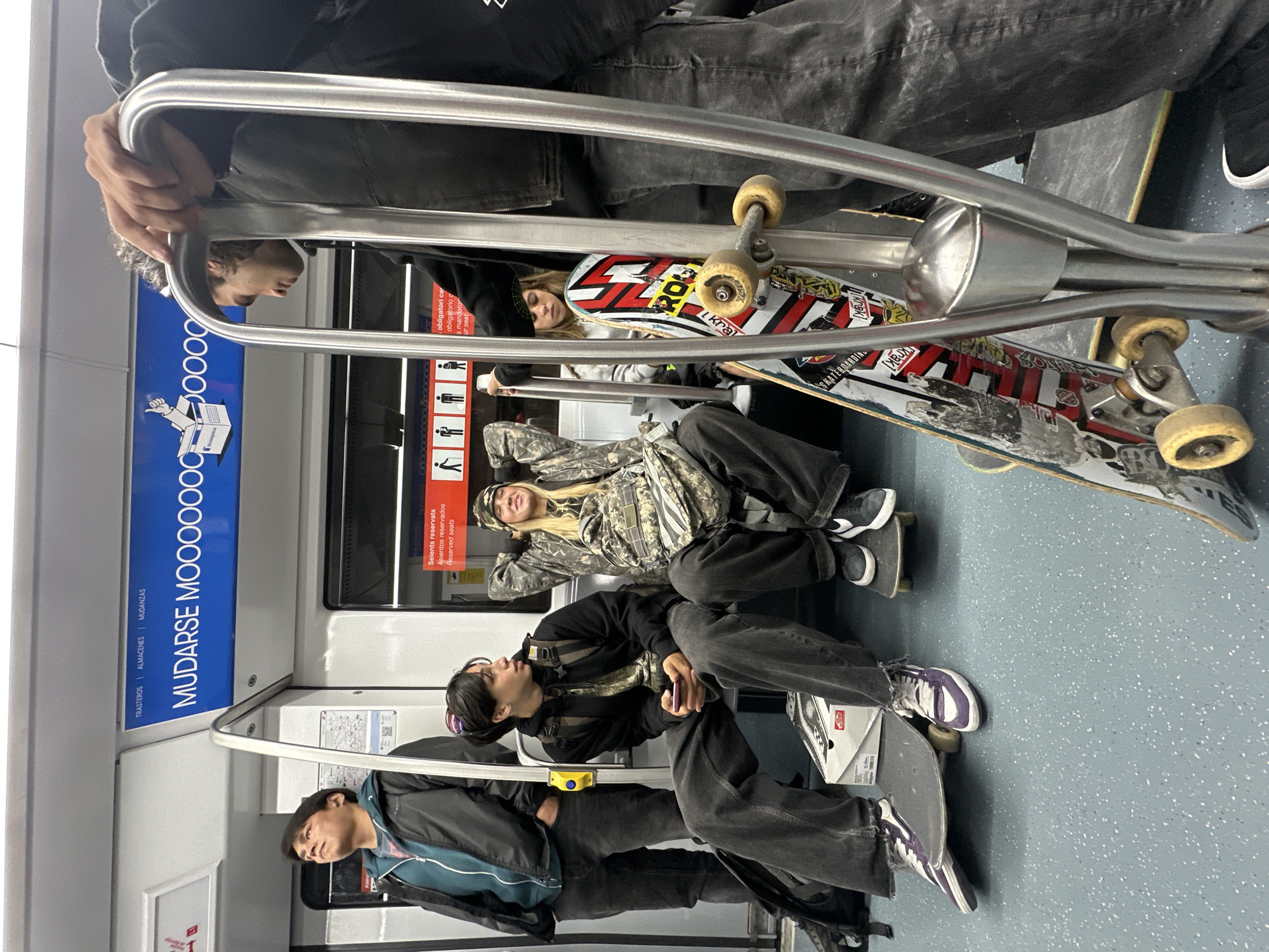 Here is a forensic description of the image:
**Scene:**
The image appears to be taken inside a public transportation vehicle, most likely a train or subway car. A blue sign with white lettering is partially visible that shows the text "TRASTEROS | ALMACENES | MUDANZAS" above and "MUDARSE MOOO0000" below and a box-moving icon, and another smaller sign is partially visible with the text "Seients reservats/Asientos reservados/Reserved seats" above "obligatori cer/obligatorio c/s mandator/ur seat" next to icons of a person seated (pregnant, elderly, handicapped) and standing with a walking stick. A metal handrail runs horizontally across the frame. The interior is generally white and grey. The floor appears to be blue with light-colored flecks.
**People:**
There are at least four young people visible in the image. All appear to be riding the train with skateboards.
* **Person 1:** At the bottom of the image, wearing a dark jacket with a teal inset.
* **Person 2:** Wearing a black jacket, black pants, and purple sneakers. They are holding what looks like a phone. They are sitting with legs outstretched, one foot resting on a skateboard.
* **Person 3:** Dressed in a camouflage patterned jacket and black pants, reclining with hands behind their head, resting on another passenger's back or shoulder.
* **Person 4:** In a light grey hoodie with a black beanie.
* **Person 5:** Arms visible on the top of the frame.
* **Person 6:** Hands visible holding the handrail at the top left of the frame.
**Objects:**
Several skateboards are present, positioned on the floor. One is propped up on top of the seat. They are stacked up, with at least four visible.
* Skateboard wheels with yellow castors
* Skateboard with white and red geometric pattern and "Kra" on the underside. Another design says "SKATEBOARDING."
**Actions/Interactions:**
The individuals seem to be traveling together, possibly heading to or from a skateboarding location. They are sitting and reclining on the train.
**Location Details**
The sign "MUDARSE" indicates a Spanish speaking area, possibly Barcelona. No other location identifiers are readily apparent.