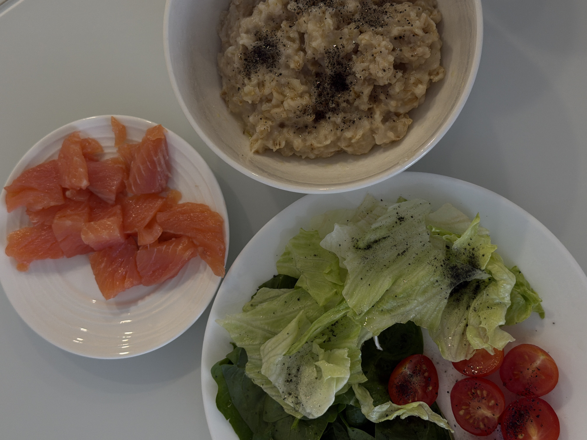 The image depicts a meal consisting of a bowl of oatmeal sprinkled with black pepper, a plate of raw salmon pieces, and a salad with lettuce, spinach, and sliced cherry tomatoes, also sprinkled with black pepper. The dishes are arranged on a white surface. There is no indication of the location being in Khimki, Russia. There are no people present in the image.