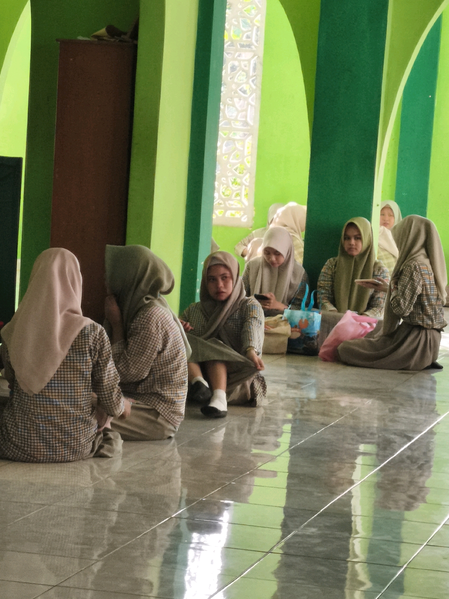 In an indoor setting with bright green walls and a tiled floor, several young girls, likely students, are gathered. They are dressed in modest clothing, including headscarves (hijabs) and long skirts or dresses. Their attire includes plaid shirts and solid-colored bottoms. The girls are seated on the floor, and their interactions suggest a moment of rest or informal gathering.
One girl in the foreground on the left is facing away from the camera, wearing a light pink hijab and a plaid shirt. Beside her, another girl, also in a plaid shirt, is turned slightly, her face partially obscured by her hand, wearing a muted green hijab. Further to the right, a girl with a green hijab is holding a smartphone. Another girl in the background, also wearing a hijab, is looking towards the camera with a neutral expression. In the far right, a girl in a brown hijab is seated with her back mostly to the camera.
Objects in the scene include two bags, one blue with a cartoon character and another lighter colored one, placed near the girls. A pink plastic bag is also visible. The environment appears to be a communal indoor space, possibly a school or a religious institution. The green wall has decorative window-like elements that are partially obscured. The tiled floor is reflective, showing the green walls and the seated figures as reflections.
The context of the city of Makassar, Indonesia, is not directly evident from the visual cues in the image alone. However, the attire of the individuals suggests a cultural context where modest dress is common, which aligns with the demographics of Indonesia.
