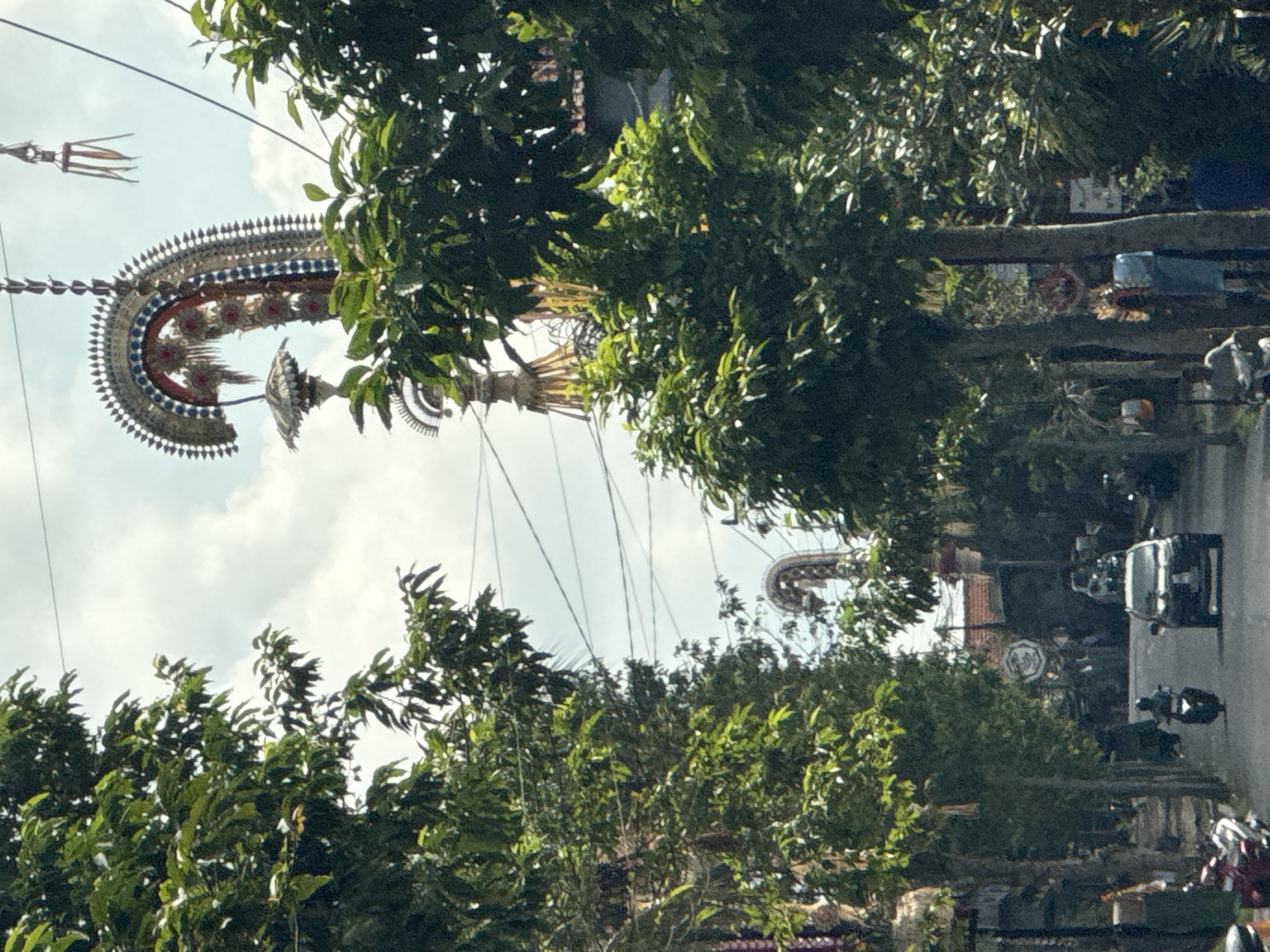 In Bangli, Indonesia, a street is lined with lush green trees, casting shadows on the asphalt road. Overhead, elaborate decorations, possibly for a festival, are suspended from wires, with one prominent, ornate structure featuring intricate patterns and hanging elements. A white car drives down the center of the road, with a person on a motorbike visible in front of it. A few other individuals are seen along the sides of the street. The sky above is a mix of white clouds and blue.