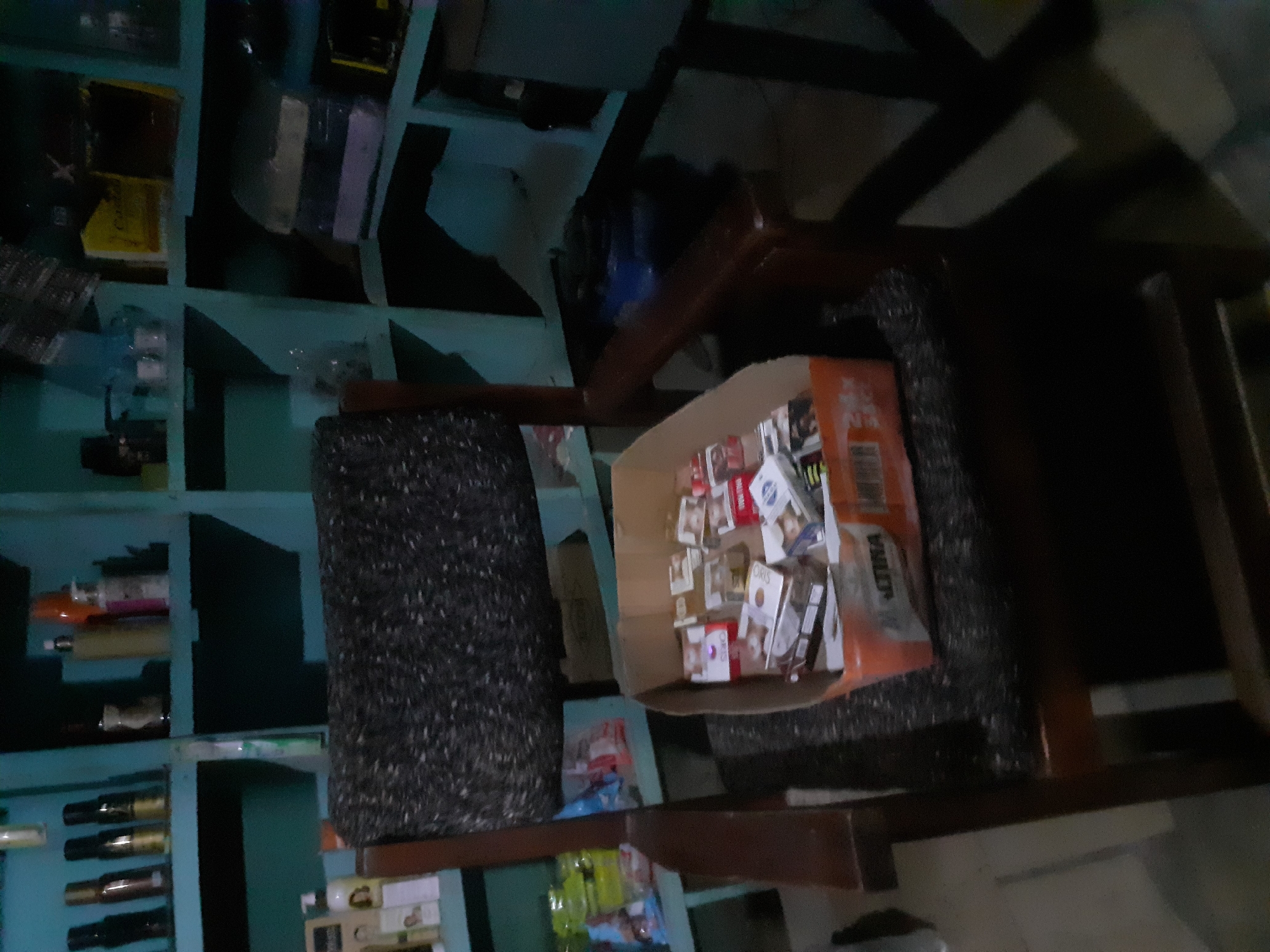 The image depicts an interior scene, possibly a room or shop, with shelves filled with various items. A wooden chair with a patterned upholstered seat and back is positioned in the foreground. A cardboard box filled with cigarette packs sits on the chair's seat. The shelves in the background contain bottles, boxes, and other packaged goods. The lighting is dim, creating shadows throughout the scene. The location is identified as Jalingo, Nigeria.