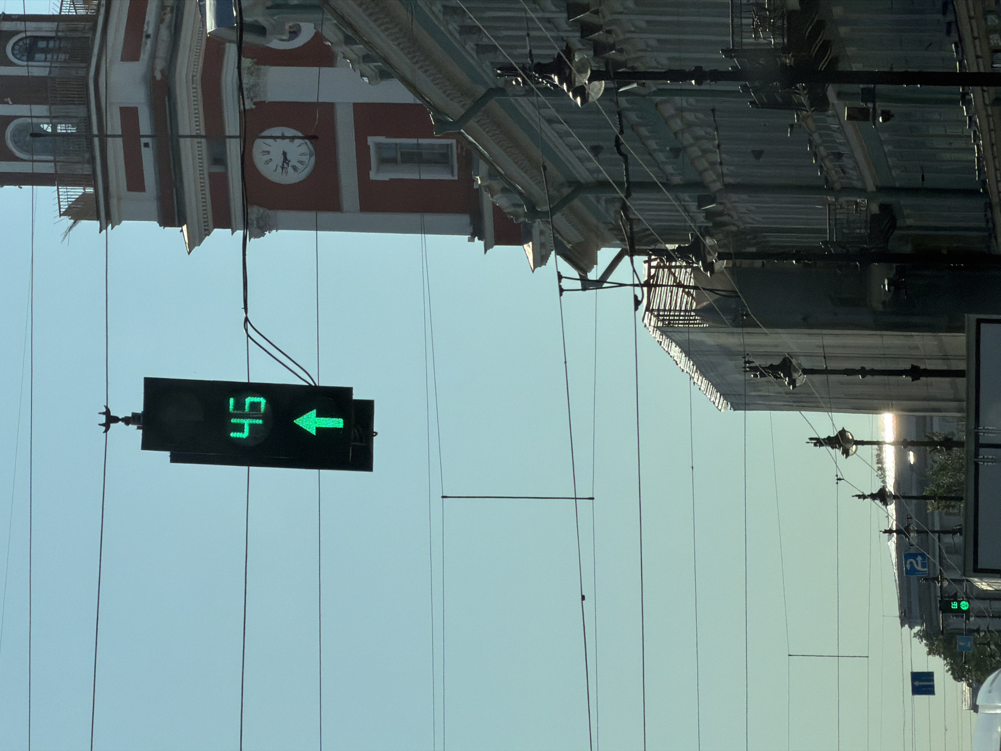 This is a medium-wide shot of an urban street scene in Saint Petersburg, Russia, on a clear day with a bright blue sky. In the foreground, a traffic light with a green arrow and the number "46" illuminated is suspended over the street by wires. To the right, a tall, red and white clock tower with arched windows and a balustrade is visible, part of an ornate building. Below the clock tower, a multi-story building with numerous windows and balconies runs along the street. Several street lamps are mounted on poles along the sidewalk. The street is spanned by a network of overhead wires, possibly for trams or trolleys. In the background, other buildings and trees are visible, with more overhead wires and traffic signs. The scene appears to be a public square or a major thoroughfare.