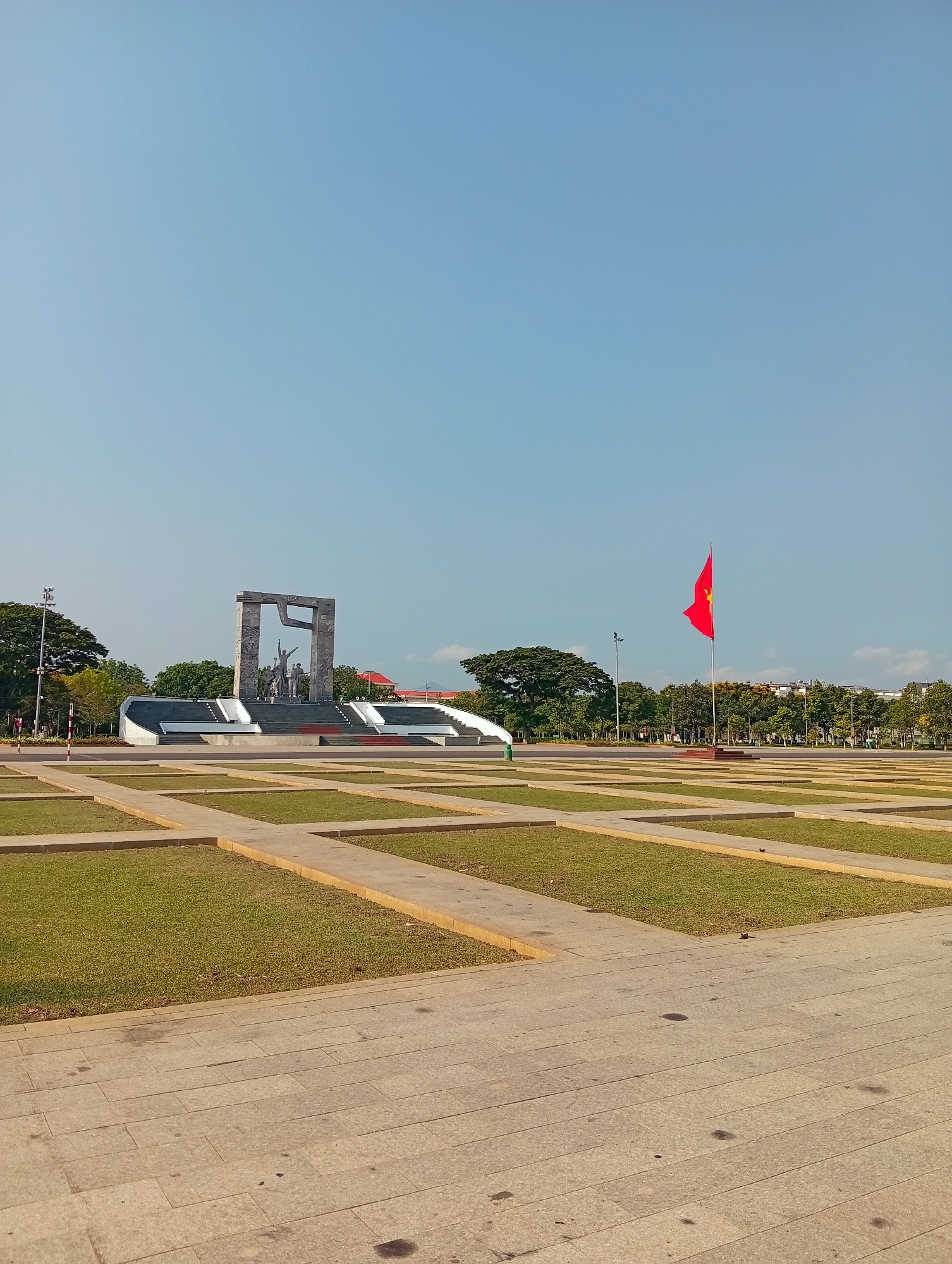 This image captures a vast, open public square in Phan Rang – Tháp Chàm, Vietnam, under a bright, clear blue sky. The scene is dominated by a large, commemorative monument in the midground, featuring a group of statues depicting human figures beneath a massive, rectangular, arch-like structure. The monument is situated on a raised, multi-tiered platform with stairs leading up to it, rendered in grey and white tones.
To the right of the monument, a vibrant red Vietnamese flag with a yellow star proudly flies from a tall flagpole, adding a striking burst of color against the blue sky. The foreground and midground of the square consist of a grid-like pattern of wide, light-colored paved paths interspersed with rectangular sections of green grass, creating a neat, geometric aesthetic.
In the distance, a dense line of trees forms a natural border for the square, with glimpses of buildings, including one with a distinctive red roof, visible through and above the foliage. A few streetlights or lampposts are also discernible near the trees. The square appears empty, with no people visible, contributing to a sense of spaciousness and quiet solemnity. The bright, even lighting indicates it is daytime, likely a sunny morning or late afternoon. No visible text is present in the image.