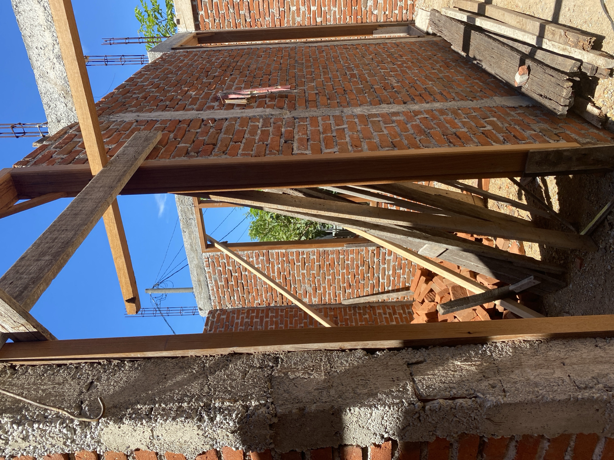 The image shows the construction of a building with brick walls and wooden beams against a clear blue sky. The walls are made of red bricks and are partially constructed, with exposed mortar. Wooden planks are visible, some supporting the structure and others stacked on the ground. There are piles of bricks near the foundation. The scene depicts an outdoor construction site in what appears to be a sunny climate.