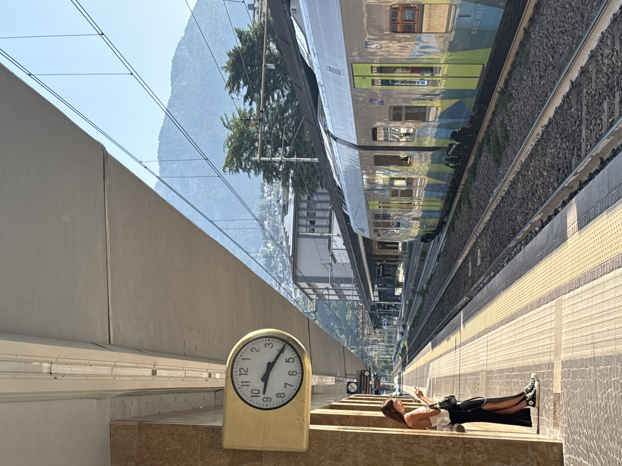 A woman stands on a train station platform in Trento, Italy, looking at her phone. She is wearing a white top and a long black skirt with a slit up the side, and black Converse sneakers. The platform has a yellow tactile warning strip along the edge. To her left, a yellow analog clock hangs on a wall. A train with a silver and green exterior is stopped on the tracks to her right. In the background, buildings and trees are visible on a mountainside under a clear blue sky. The station appears to be covered by a large, sloped concrete overhang.