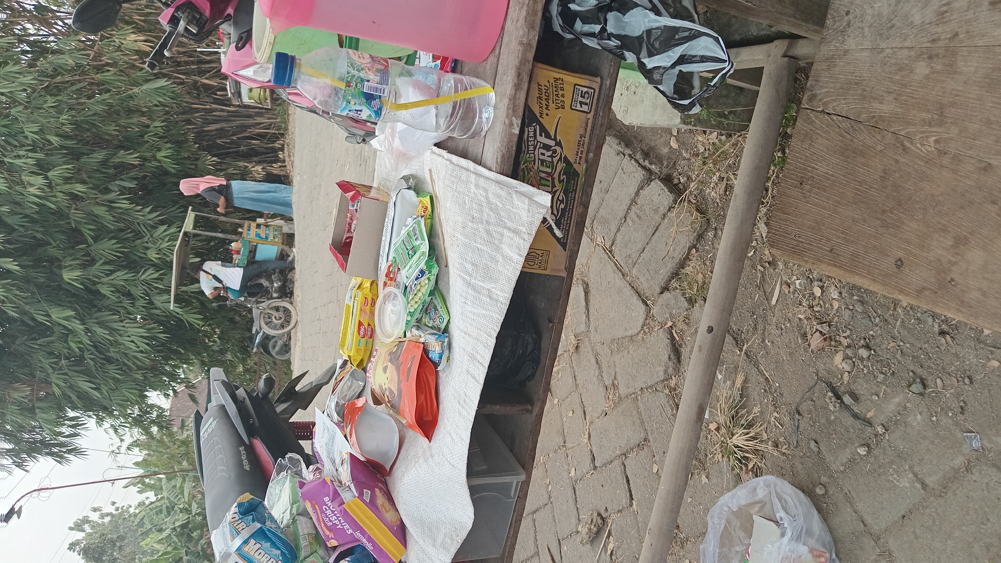 In Kedungwaru, Indonesia, a scene unfolds on a paved area, likely a roadside, with a thick bamboo grove forming a natural backdrop. The foreground is dominated by a low wooden table laden with an assortment of items. Among them are various packaged snacks, including what appear to be wafers and candy, along with an open red box. A plastic bottle of water with a yellow straw stands prominently, next to a pink pitcher. Underneath the table, a black plastic bag is visible, along with a cardboard box bearing "HALAL" and "GINSENG."
Behind the table, a food cart with a white canopy is situated, attended by two individuals. One, wearing a white t-shirt, stands behind the cart, seemingly engaged in preparing or serving food. The other, a person in a light pink hijab and a blue skirt, stands facing the cart, possibly a customer. A motorcycle is parked next to the cart, and another motorcycle is partially visible to the left of the frame. The ground is a mix of paving stones and dirt, with dry grass and scattered debris. A rough wooden plank lies in the lower right portion of the image, and a white plastic bag filled with various items is near the left edge. The overall impression is of a casual, perhaps slightly disheveled, roadside vending area.