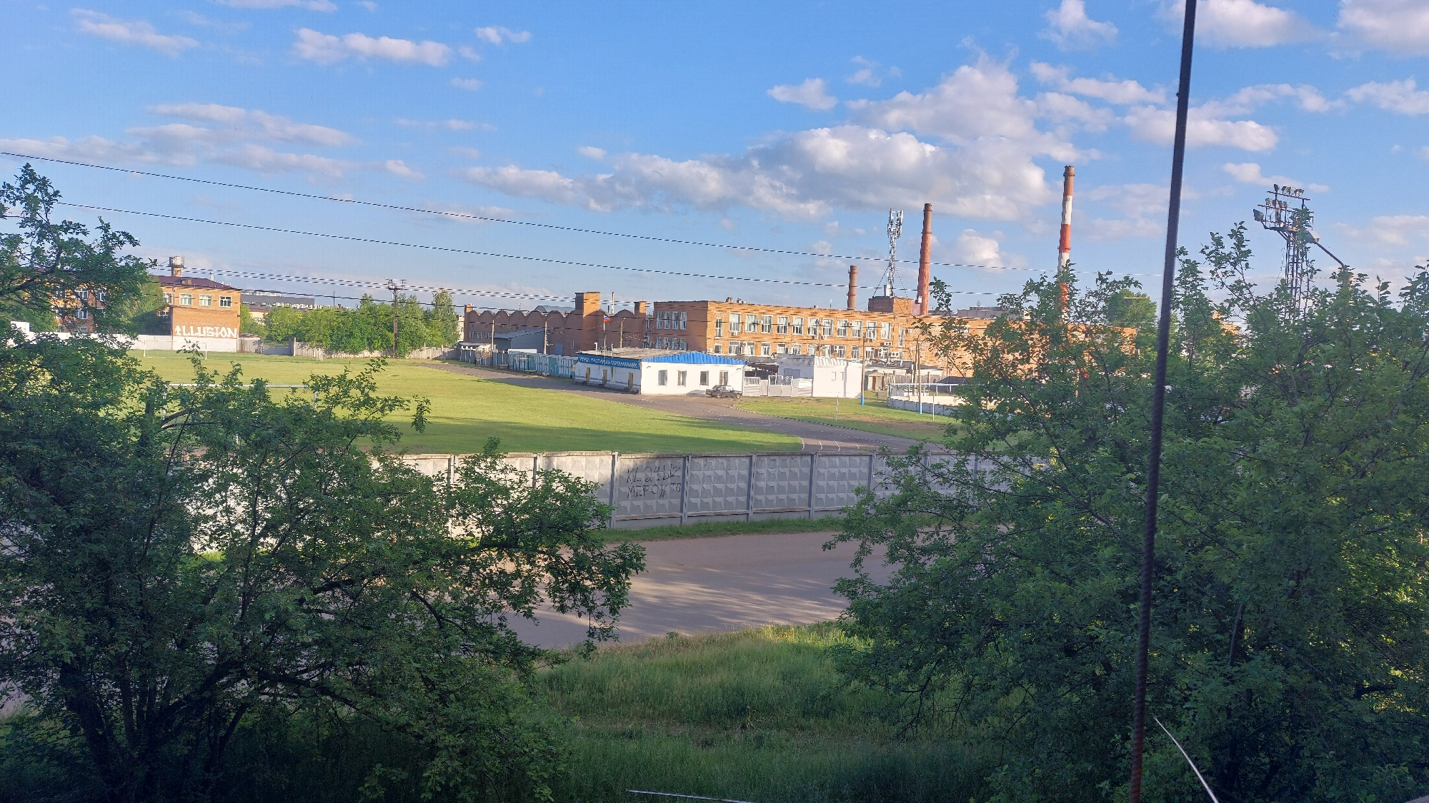 The image shows a daytime view of an industrial area with factories and smokestacks under a blue sky with scattered clouds. In the foreground, lush green trees partially obscure the view. A concrete fence with a patterned surface runs horizontally across the middle ground, with some graffiti visible on it. Beyond the fence, a large, open grassy field is present. In the background, several brick industrial buildings are visible, some with multiple stories and windows. Two tall, red and white striped smokestacks stand prominently against the sky, along with a transmission tower equipped with antennas. A dark-colored sedan is parked on a paved road near one of the buildings. The overall impression is that of an operational industrial zone on a clear day. The provided information indicates the location is in Kansk, Russia.