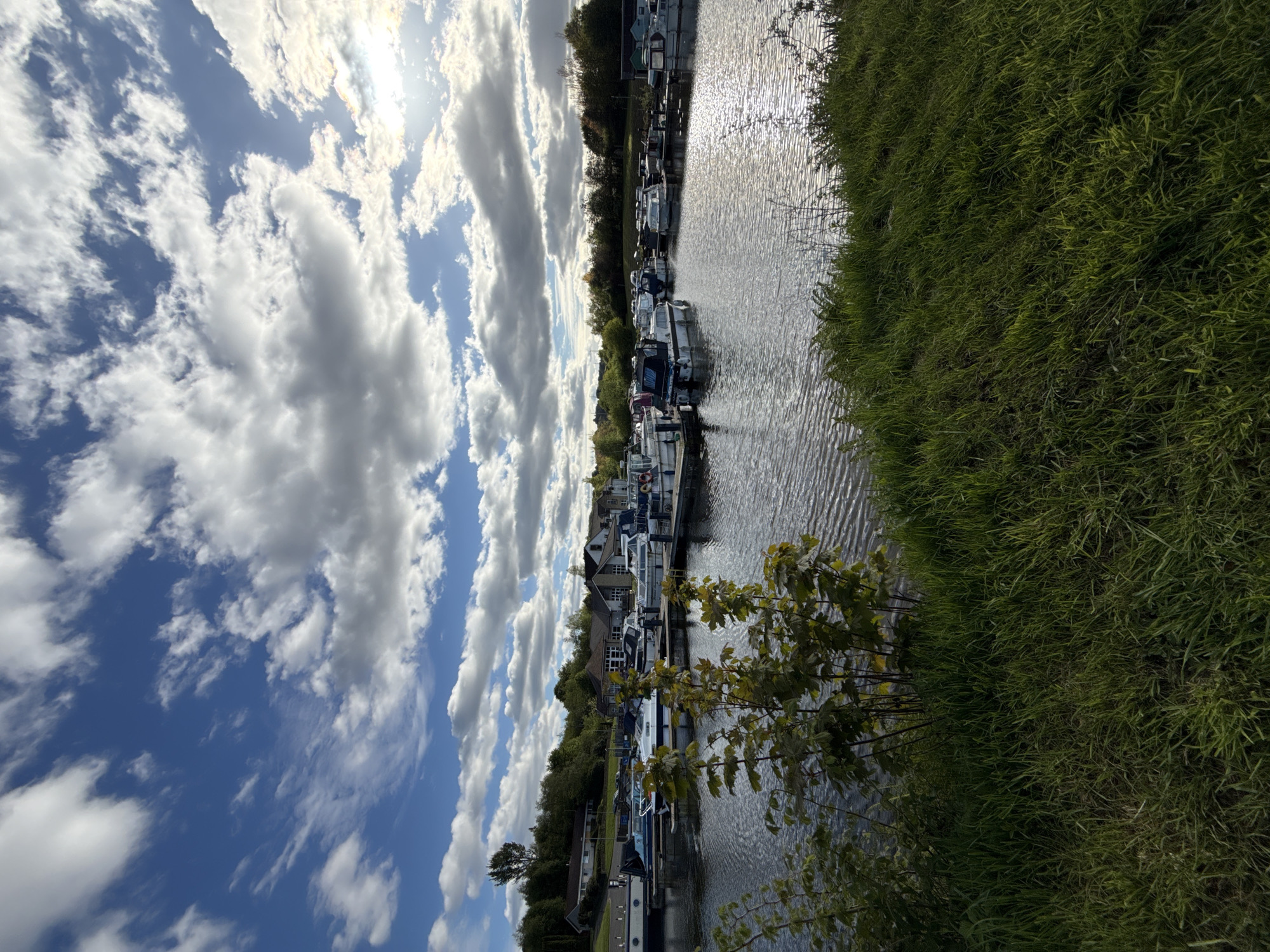 The image shows a canal scene with moored boats under a sky with plentiful clouds. The camera appears to be positioned from the bank of the canal. Lush green grass covers the bank on the right. The canal is filled with water that reflects the light, creating a shimmering effect. Moored boats are lined up on the opposite side of the canal, docked closely together. Behind the boats are buildings and trees. The sky is a vivid blue with white clouds. The location is Unknown, Unknown.