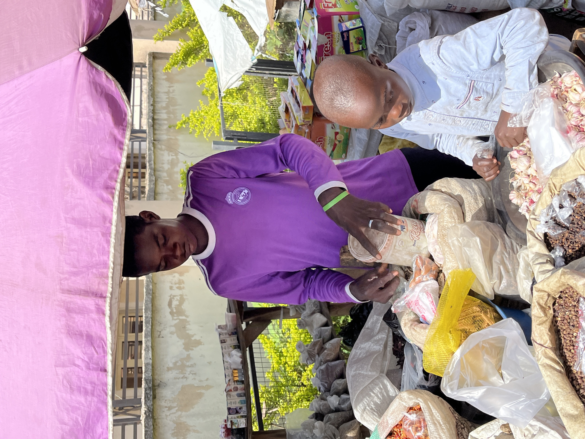 Two vendors are working at a market stall in Jos, Nigeria. A man in a purple Real Madrid jersey is pouring from a container into a package. A boy in a white shirt is handling items at the stall, which is filled with various food items and spices packaged in bags. A pink umbrella covers part of the stall. Boxes of goods are in the background. There is an exterior building behind the stall.