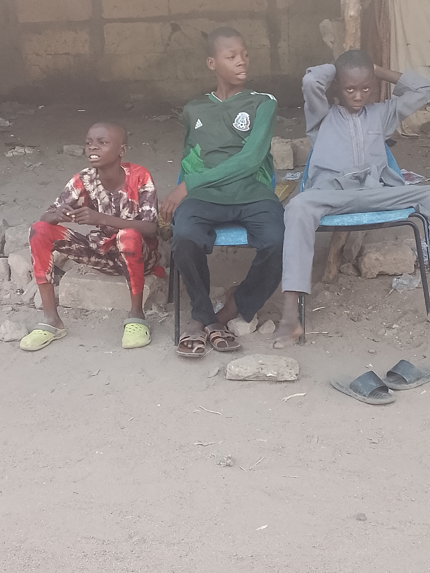 Three boys are sitting outside a building. One is sitting on a low stone, wearing a multi-colored shirt and pants with yellow Crocs. The second boy sits on a blue chair, wearing a green soccer jersey and black pants with brown sandals. The third boy sits on a second blue chair, wearing a grey robe with bare feet. A pair of dark-colored sandals is on the ground. The scene appears to be in a dusty area in Pantame, Nigeria.