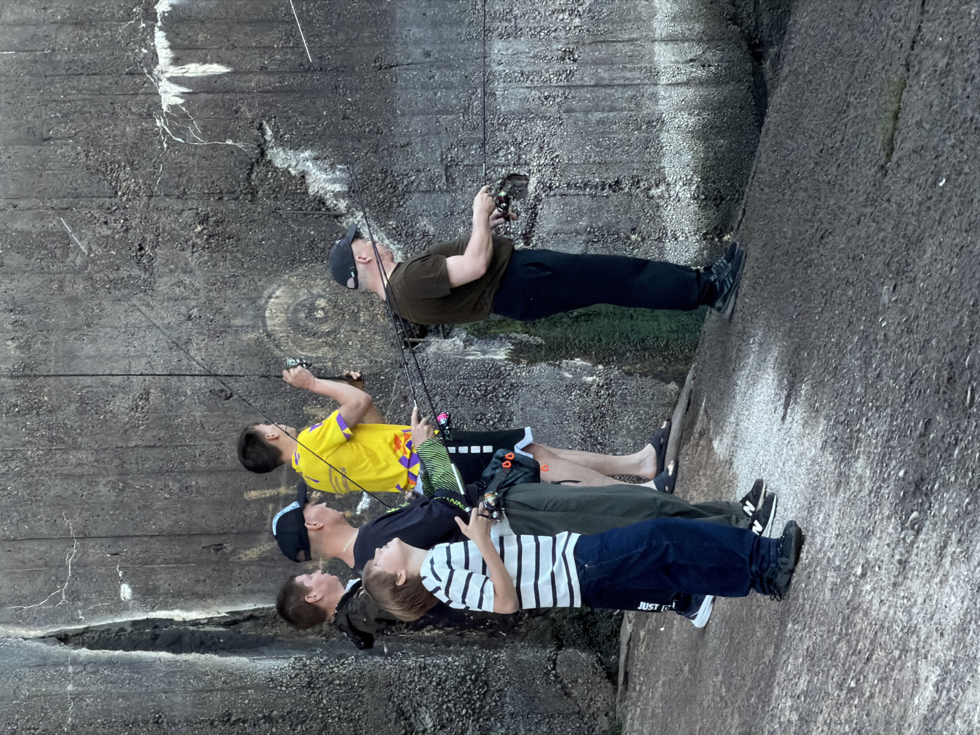 A group of five individuals, including at least one juvenile, are fishing near a large, weathered concrete structure, possibly a dam or bunker. The concrete wall shows signs of age and damage, with cracks and discolored patches. The ground beneath them is rough and uneven, a mix of rock and gravel. The individuals are positioned at various points along the wall, each holding a fishing rod.
The person furthest to the left, a young male, wears a camouflage-patterned jacket and dark pants. Next to him, a man in a black t-shirt and a black baseball cap is also fishing. A young boy in a striped t-shirt and dark pants stands in front of them, looking upwards. To the right of the boy, a person in a bright yellow t-shirt with purple accents is fishing. The furthest person to the right, wearing a dark t-shirt and a black baseball cap, is also engaged in fishing. The overall scene suggests a casual outdoor activity, with the imposing concrete structure providing a stark backdrop. The location is identified as being in the city of Chaikovsky, Russia.