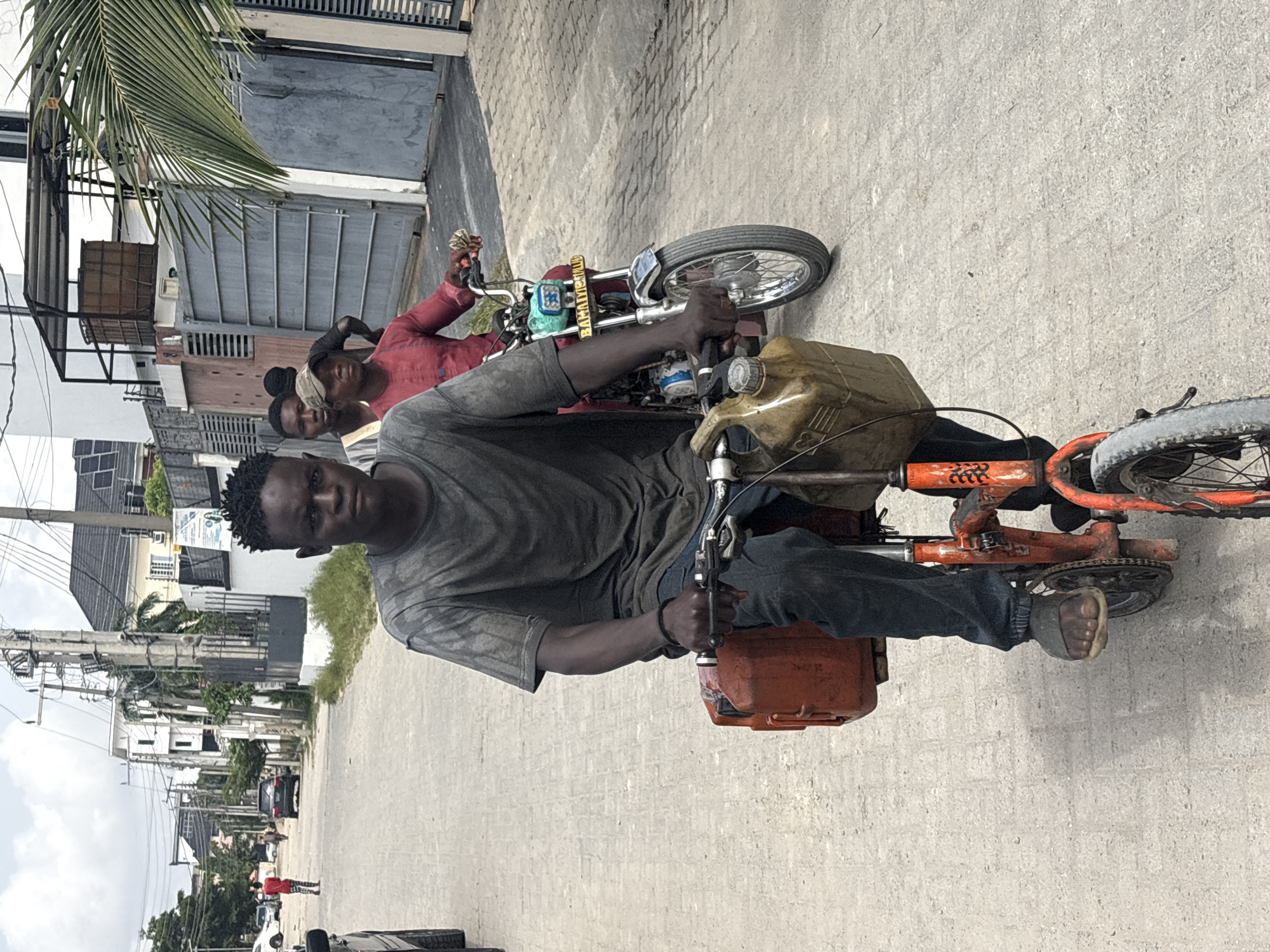 A man wearing a gray t-shirt and jeans is riding a modified orange bicycle on a paved street in Igboefon, Nigeria. He is barefoot. Attached to the bicycle are two large plastic containers, one orange in the front and a tan color in the back. Another man in a red shirt is standing next to a red motorcycle with a license plate nearby. Additional people and buildings line the street.