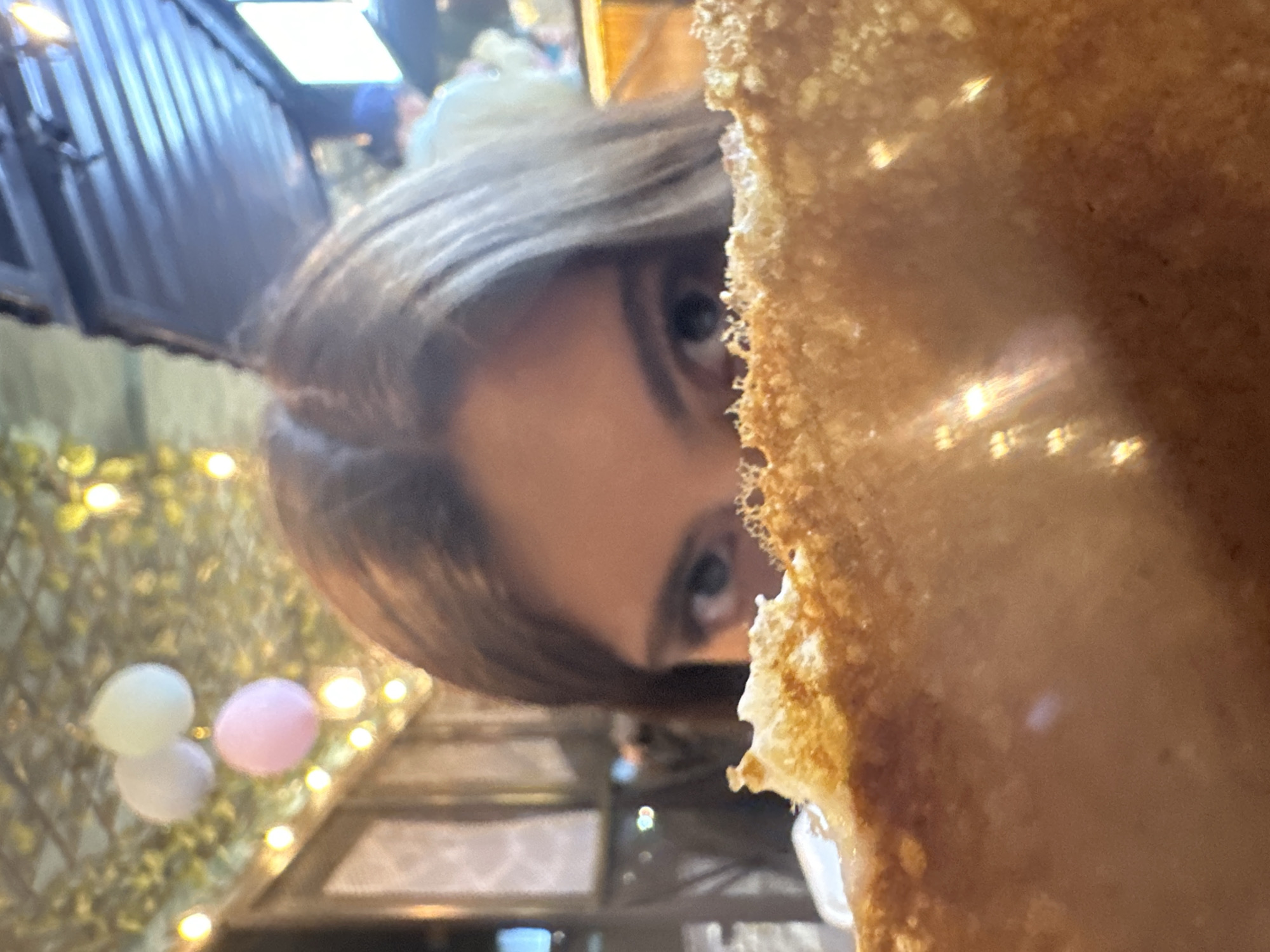 The image shows a close-up of a person's face partially obscured by what appears to be a large, crispy food item, likely a crepe or similar pastry. The person has brown hair and visible eyes, with the focus directed towards them. The background shows a blurred setting of an outdoor restaurant. Based on the visual cues, the location is likely Puebla, Mexico, given the commonality of street food like crepes and the presence of decorative lights and balloons, suggesting a festive or dining atmosphere often found in Puebla's city center or tourist areas. The exact address or building cannot be determined from this image.