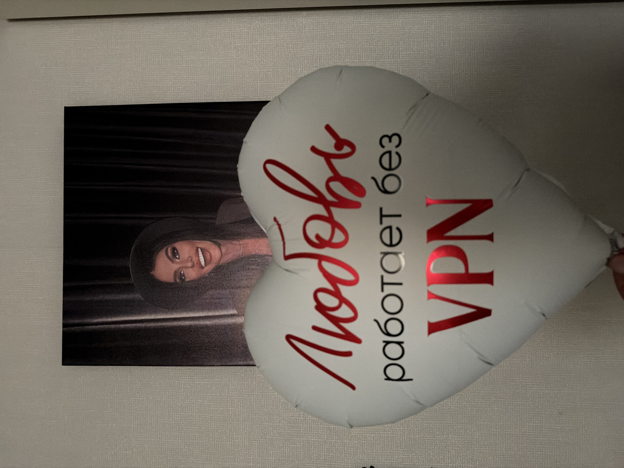 The image shows a white heart-shaped balloon with the text "Любовь работает без VPN" in Russian, meaning "Love works without VPN," written in red and black. The balloon is partially obscuring a framed portrait of a woman with dark hair and a wide-brimmed hat. The background is a neutral-toned wall. The location is likely Moscow, Russia, due to the Cyrillic text and associated VPN usage messaging, often tied to censorship concerns within the country.