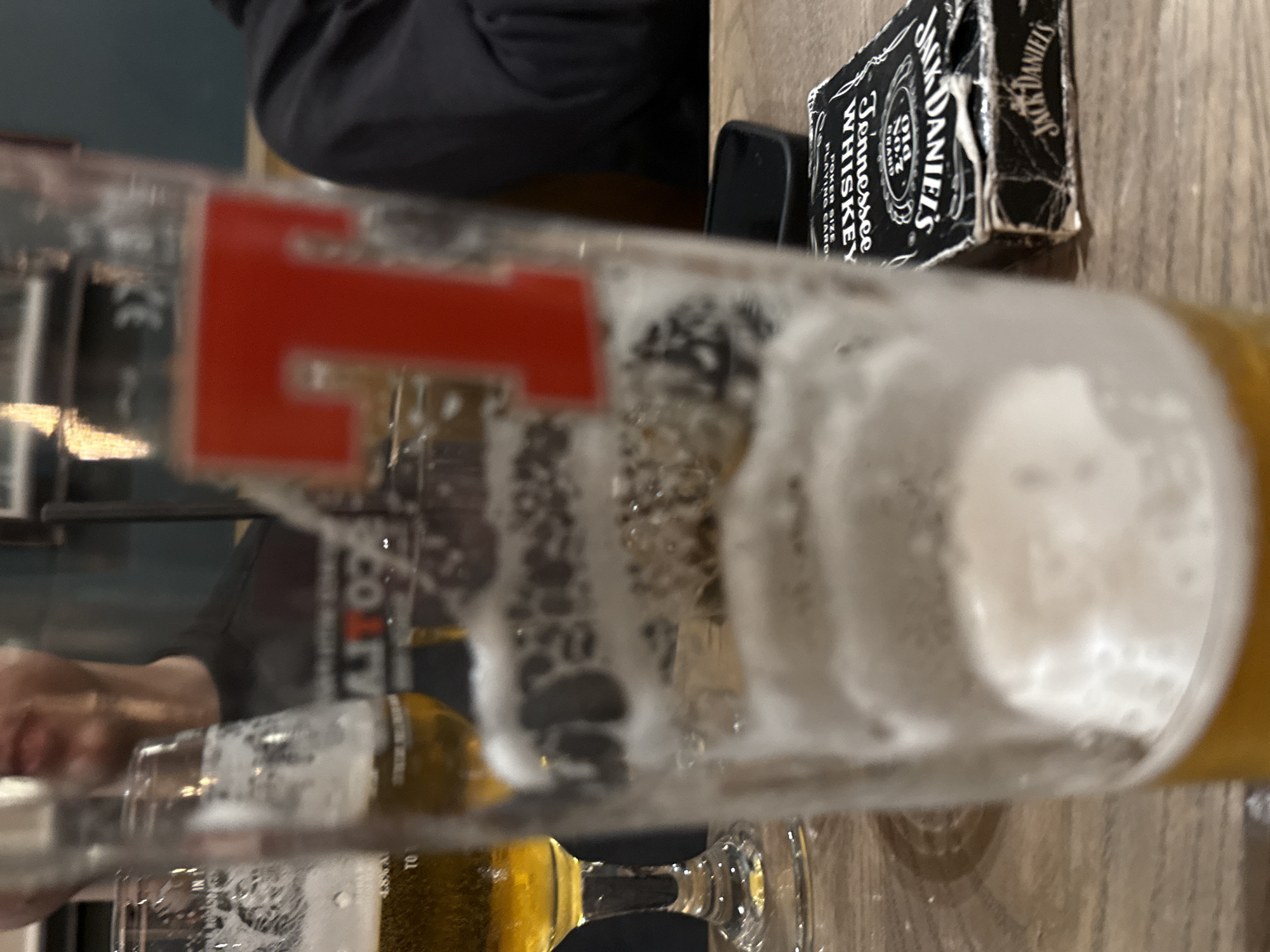 The image shows a close-up of a partially filled beer glass with a red "T" logo on it. The glass is on a wooden table. Next to the glass is a deck of Jack Daniel's Tennessee Whiskey playing cards. A mobile phone is also partially visible near the playing cards. A person is seen blurred in the background on the left side of the image. The image gives the impression that the location is a bar or a pub.