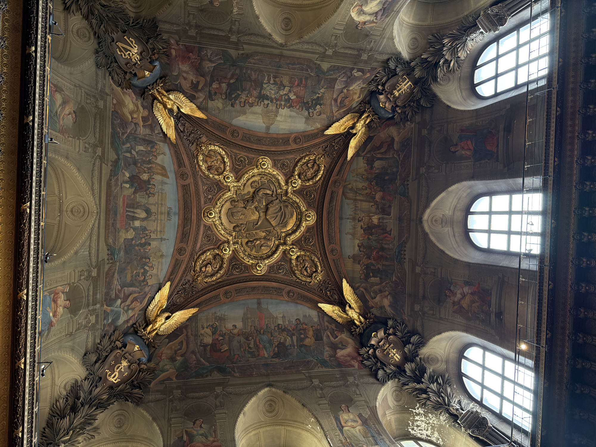The image depicts the painted ceiling of the Salle des Etats in the Louvre Museum, Paris, France. The ceiling is ornately decorated with paintings depicting historical scenes and allegorical figures. Gold eagles are positioned at the cardinal points of the central decorated section, and are flanked by decorative elements including laurel and shields. Natural light enters the room from tall arched windows visible on the right side of the image. A maintenance structure with lights is positioned in front of the windows. The paintings portray large groups of people.