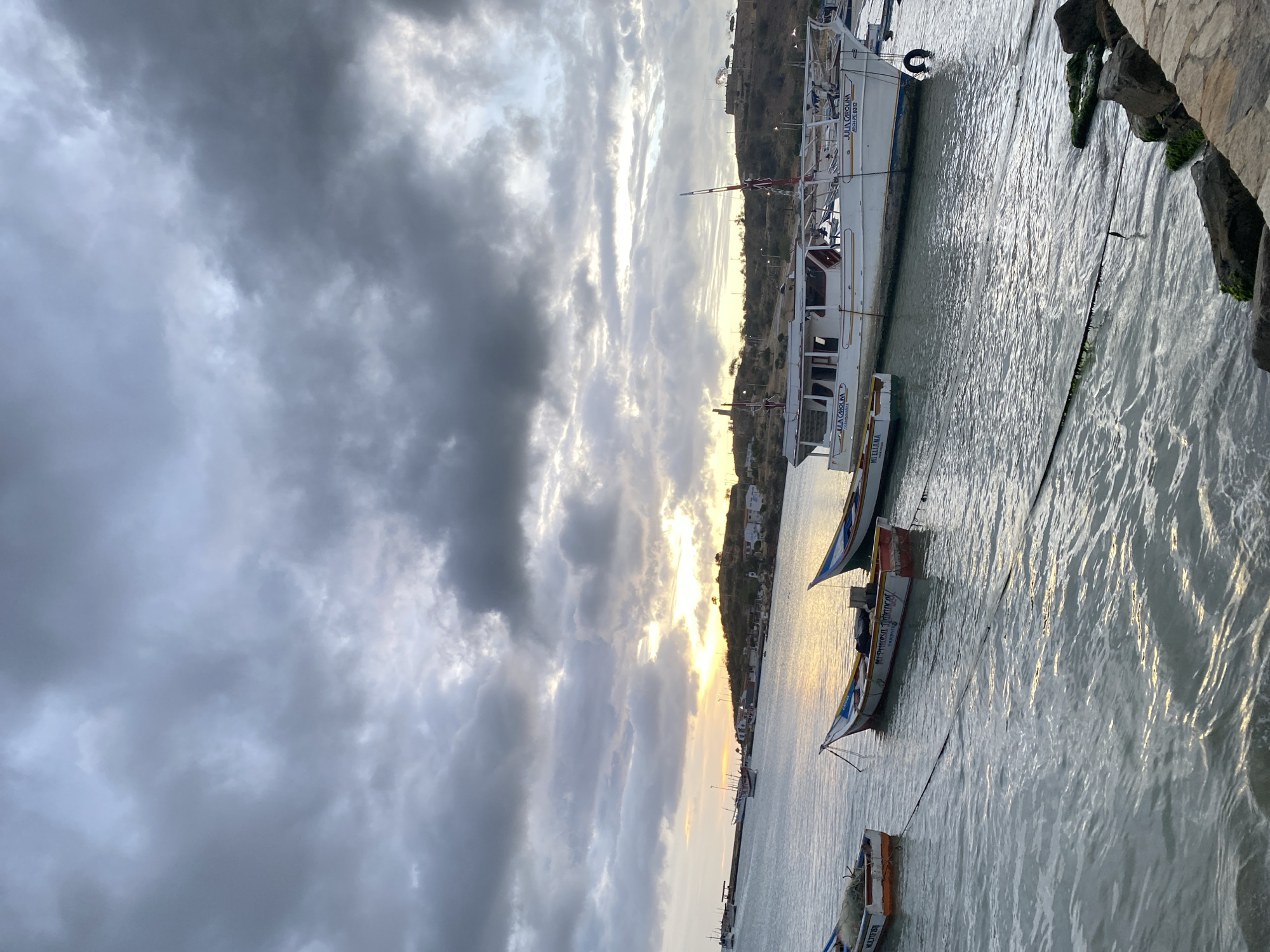 The image shows a harbor scene in Juan Griego, Venezuela. A large white boat is docked alongside a concrete pier covered with algae and moss. Two smaller boats are moored nearby. The water is choppy, with sunlight reflecting off the surface. The sky is overcast with heavy clouds, though a strip of sunlight is visible on the horizon. Buildings are visible on the hillside in the background.