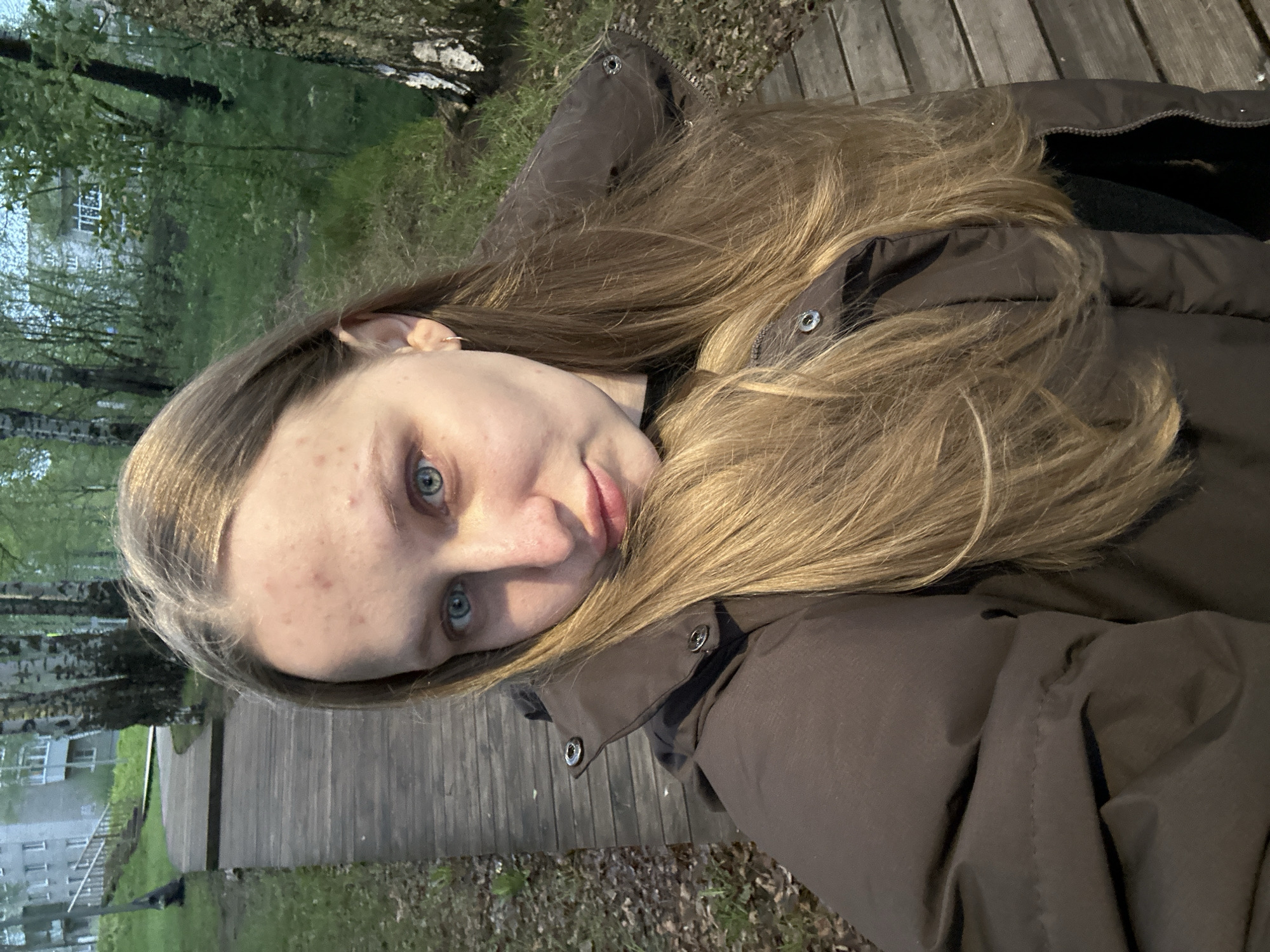 The image shows a young woman with long blonde hair taking a selfie outdoors. She is wearing a brown puffer jacket. The background consists of trees, grass, a wooden walkway, and a building in the distance. The location appears to be a park or green space in the city of Chaikovsky, Russia.