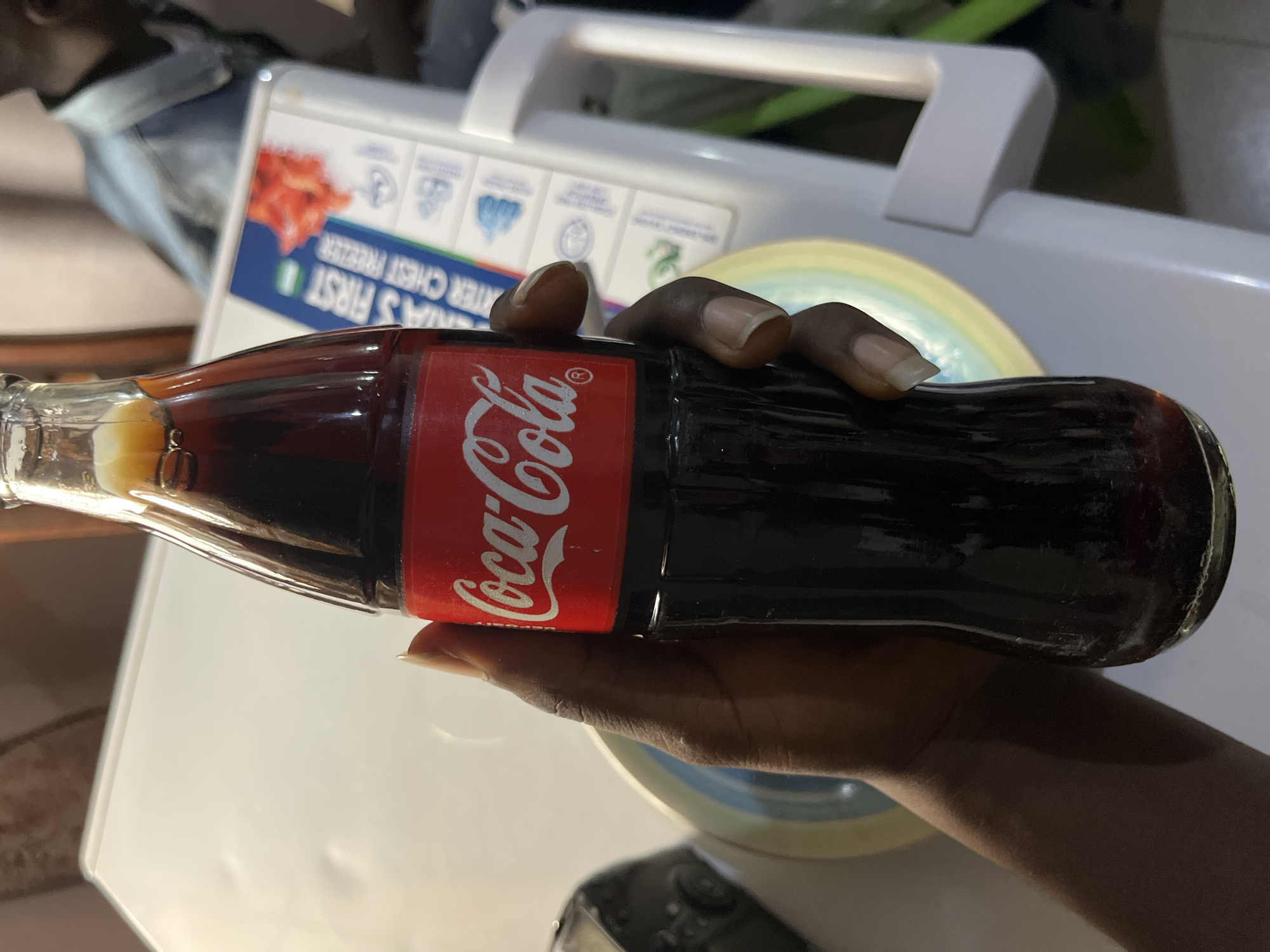 A person's hand, with dark skin and neatly manicured fingernails, holds a glass bottle of Coca-Cola. The bottle is mostly full of dark brown liquid and has the iconic red and white Coca-Cola label. The background appears to be a kitchen or dining area, with a white surface (possibly a table or refrigerator) visible. There is a glimpse of a person wearing a denim jacket in the far background. The scene suggests a casual, everyday moment. The image appears to have been taken indoors, likely in a residential setting in Bukuru, Nigeria.