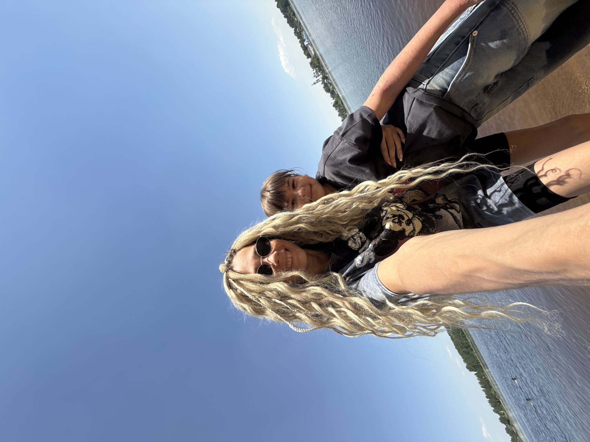 A rotated, medium shot of a woman with braided blonde hair, wearing sunglasses, and a young child outdoors in the city of Unknown, Unknown. The woman and child are on what appears to be a shore. In the background is water with some vegetation along the bank. The sky is clear and blue. The woman is smiling, and the child is looking at the camera with a small smile. The woman is holding the child, who is wearing a dark t-shirt and black shorts. Part of another person's leg can be seen.