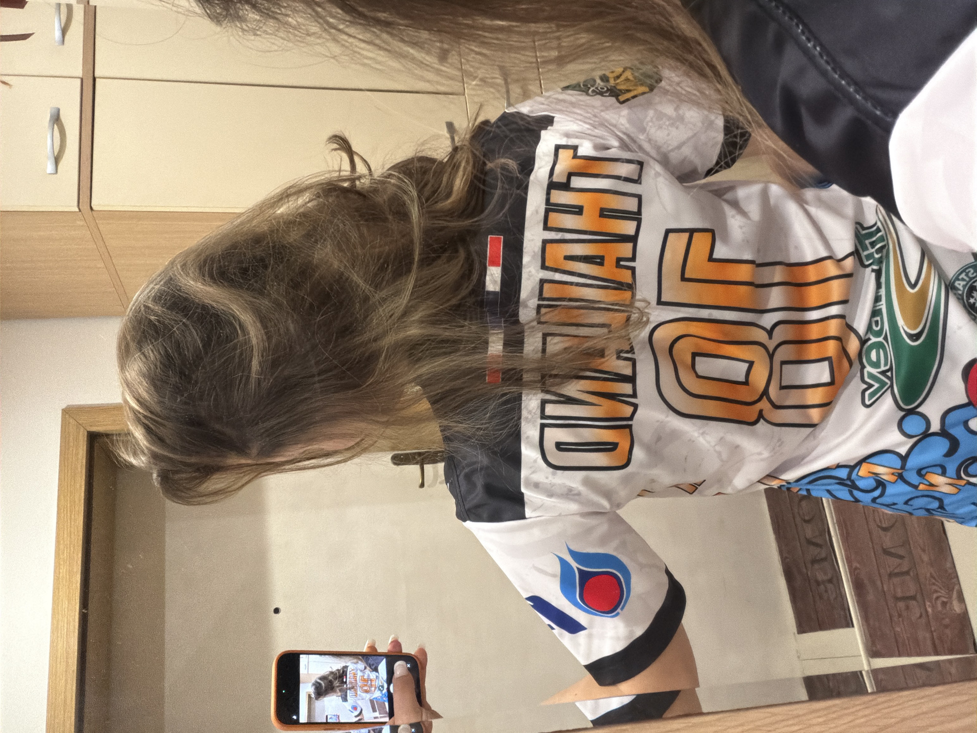 The original image is rotated 90 degrees counter-clockwise, showing a person from behind, leaning forward and to the left. The person has long, wavy light brown hair covering much of their back and shoulders. They are wearing a white sports jersey with black sleeves and colorful designs.
Prominently displayed across the upper back of the jersey is the inverted word "THAILAND" in large orange and black outlined letters. Below this, large numbers, appearing to be "86", are also visible in a similar style. A small rectangular flag-like emblem, resembling the flag of Thailand, is visible on the upper left shoulder area of the jersey. On the left sleeve, there is a logo featuring red and blue flame-like elements (likely PTT), and on the right sleeve, a green and yellow/gold crest-like logo.
The person is taking a mirror selfie, holding an orange-cased smartphone in their right hand. The phone's screen reflects a miniature, inverted image of the person's back and jersey. The person's right hand features manicured fingernails with white tips. The mirror reflects parts of an indoor room behind the person, including light-colored wooden cabinets with silver handles and a wooden door frame against plain, light-colored walls. The person's face is not visible. The scene is set indoors, in Varna, Bulgaria.
