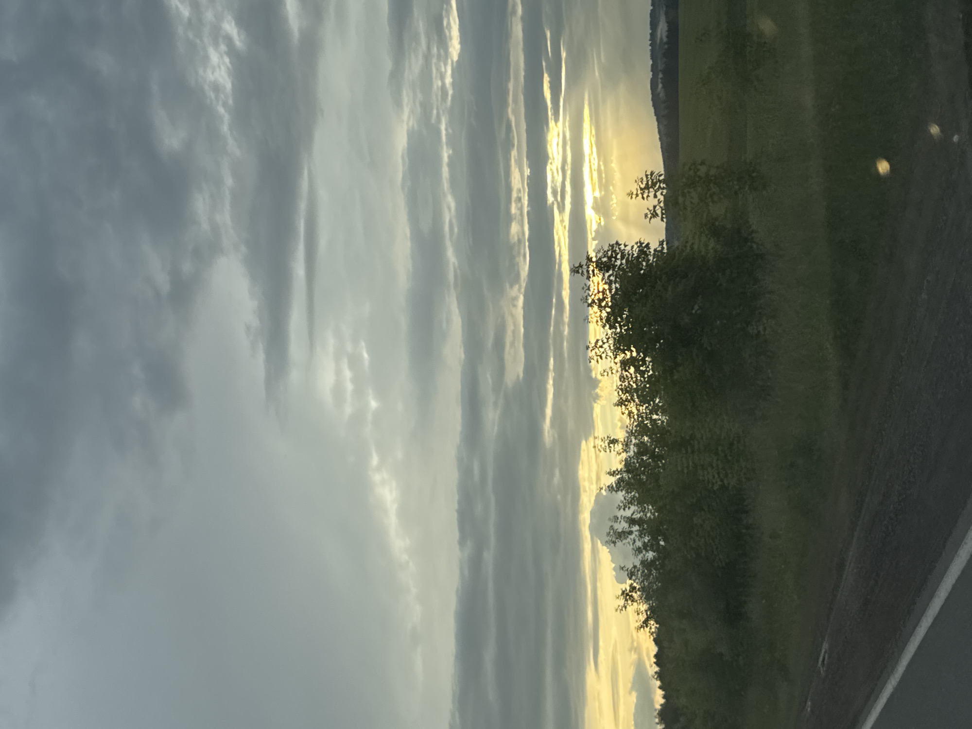 The image depicts a scenic view of a cloudy sky at dusk, with the sun's rays peeking through the clouds and casting a warm glow on the landscape. The sky is filled with dramatic clouds, ranging from dark grays to bright whites, suggesting the possibility of an approaching storm or the remnants of one. The setting appears to be a rural area in Забегалово, Russia, with a lush green field and a line of trees bordering the right side of the frame. A dark asphalt road, possibly a highway, runs along the bottom right edge of the image, with a white line marking the lane. The overall mood is one of tranquility and natural beauty, with the dramatic sky adding a touch of awe. The time of day is clearly late afternoon or early evening, given the quality of the light. There are no people or objects explicitly visible other than the natural elements.