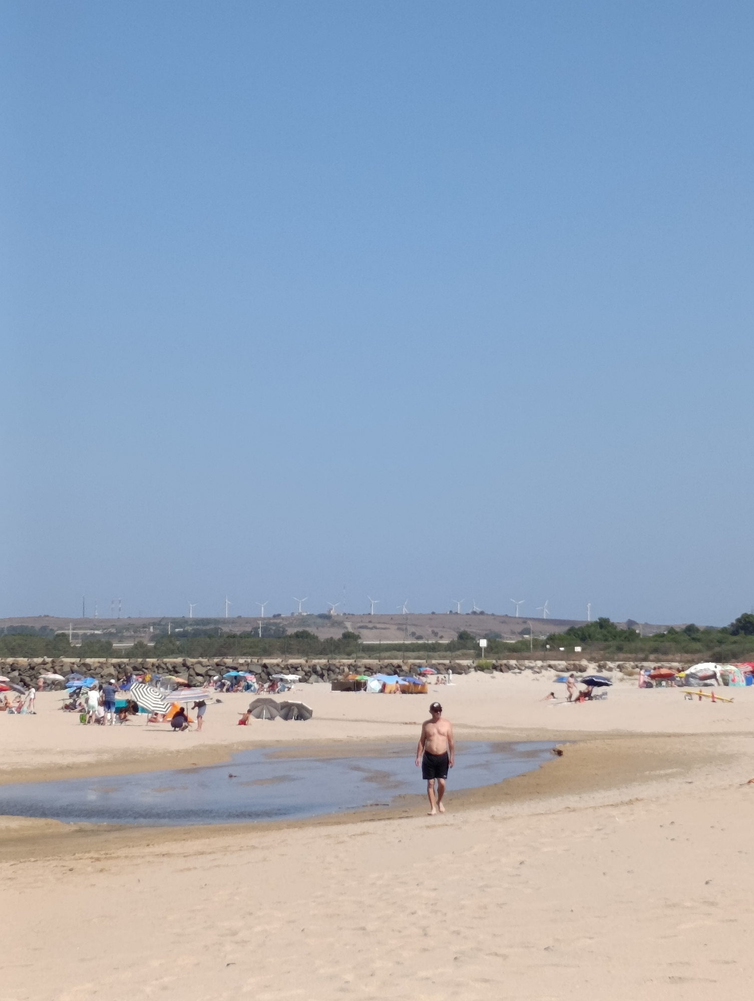 The image depicts a sunny day at a sandy beach in Sines, Portugal. In the foreground, a man wearing black shorts walks towards the viewer across the sand. Behind him, a shallow pool of water with a sandy edge separates him from the main beach area. The main beach is populated with numerous beachgoers, many of whom are sitting under colorful umbrellas. Beach tents and shelters are also visible. In the middle ground, a line of rocks forms a barrier, and beyond that, a hillside is dotted with green vegetation and several wind turbines. The sky is clear and blue.