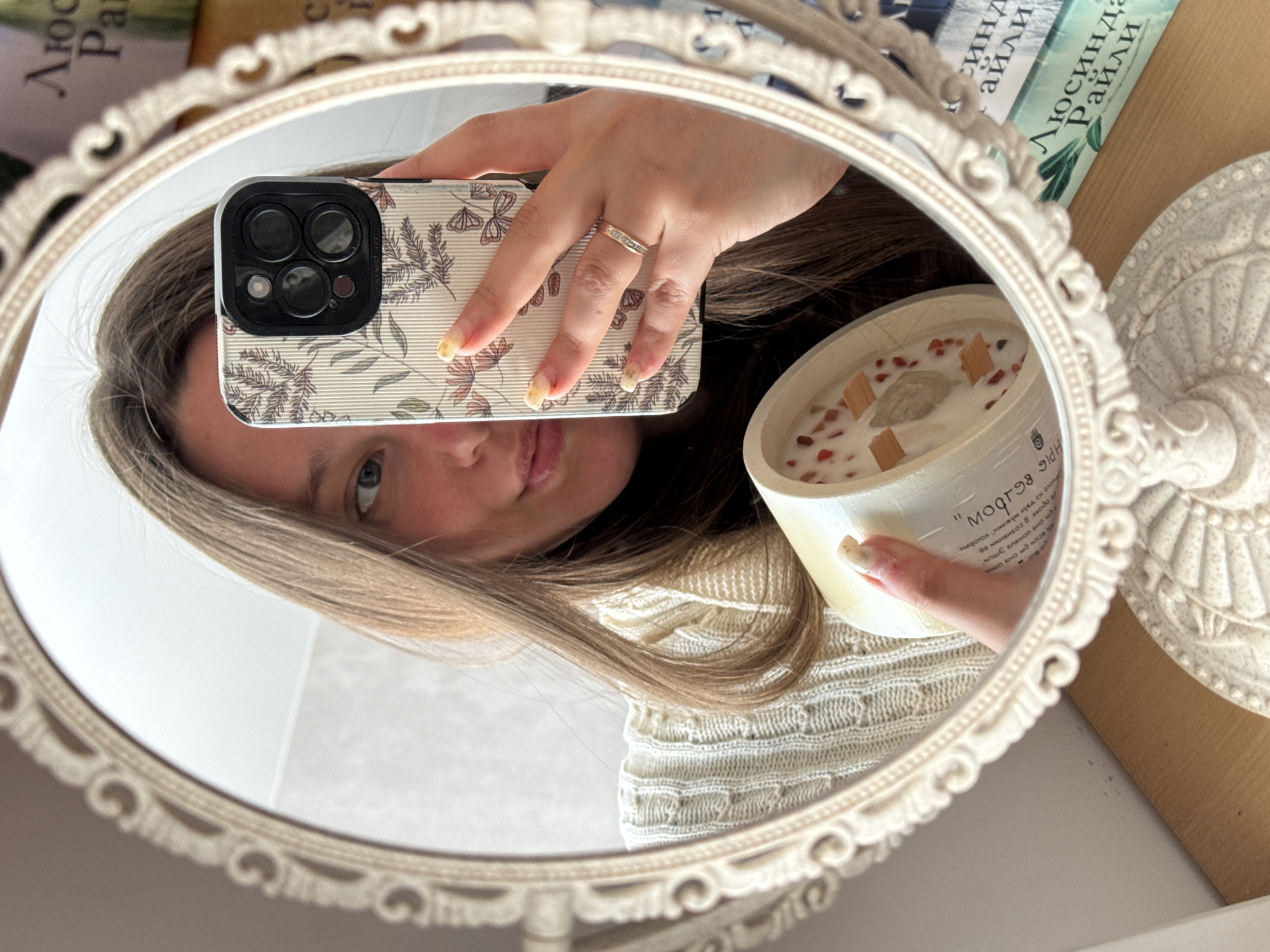 An indoor mirror selfie shows a young person holding a smartphone and a decorative candle, reflected in an ornate, oval-shaped mirror with a light-colored, scrolled frame. The subject, with long, light brown hair, is partially visible, showing one eye and her mouth, and is wearing a cream-colored, textured knitted sweater.
She holds a smartphone, likely an iPhone, in a floral-patterned case featuring botanical designs and butterflies on a ribbed background. A ring adorns one finger, and her nails are painted with a light yellowish polish showing some wear. Her other hand holds a candle in a light-colored jar, which contains three visible wooden wicks, small red/orange decorative pieces, and a larger, clear stone or crystal embedded in the wax. The candle label, mirrored, displays "МООЯН" (implying "BLOOM" forwards) and other partially visible Cyrillic text.
In the background, reflected above the mirror, are several books with Cyrillic text, including "Люсинда Райли" (Lucinda Riley) and parts of a title like "Синд" (likely from "The Seven Sisters" series). Part of a decorative, light-colored stand is visible to the right of the mirror. The scene reflects a personal space in Krasnodar, Russia. No specific time of day or weather is indicated.