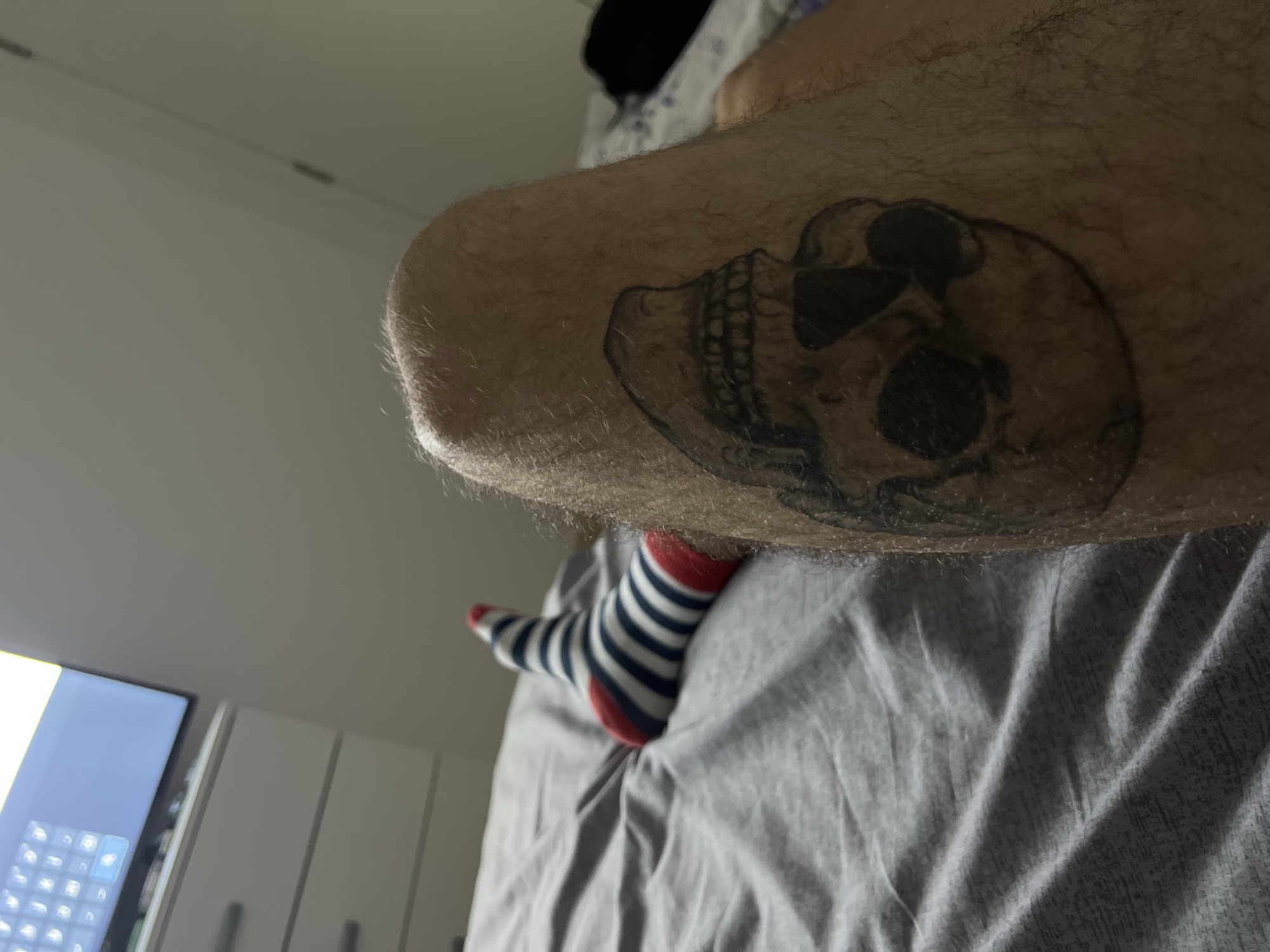 The image shows a man's leg with a skull tattoo. The leg is resting on a gray wrinkled bed sheet. The man is wearing a blue and white striped sock with a red toe and heel. In the background, a portion of a television screen and a white cabinet can be seen. The image does not contain information to determine the location in Rafailovići, Montenegro.