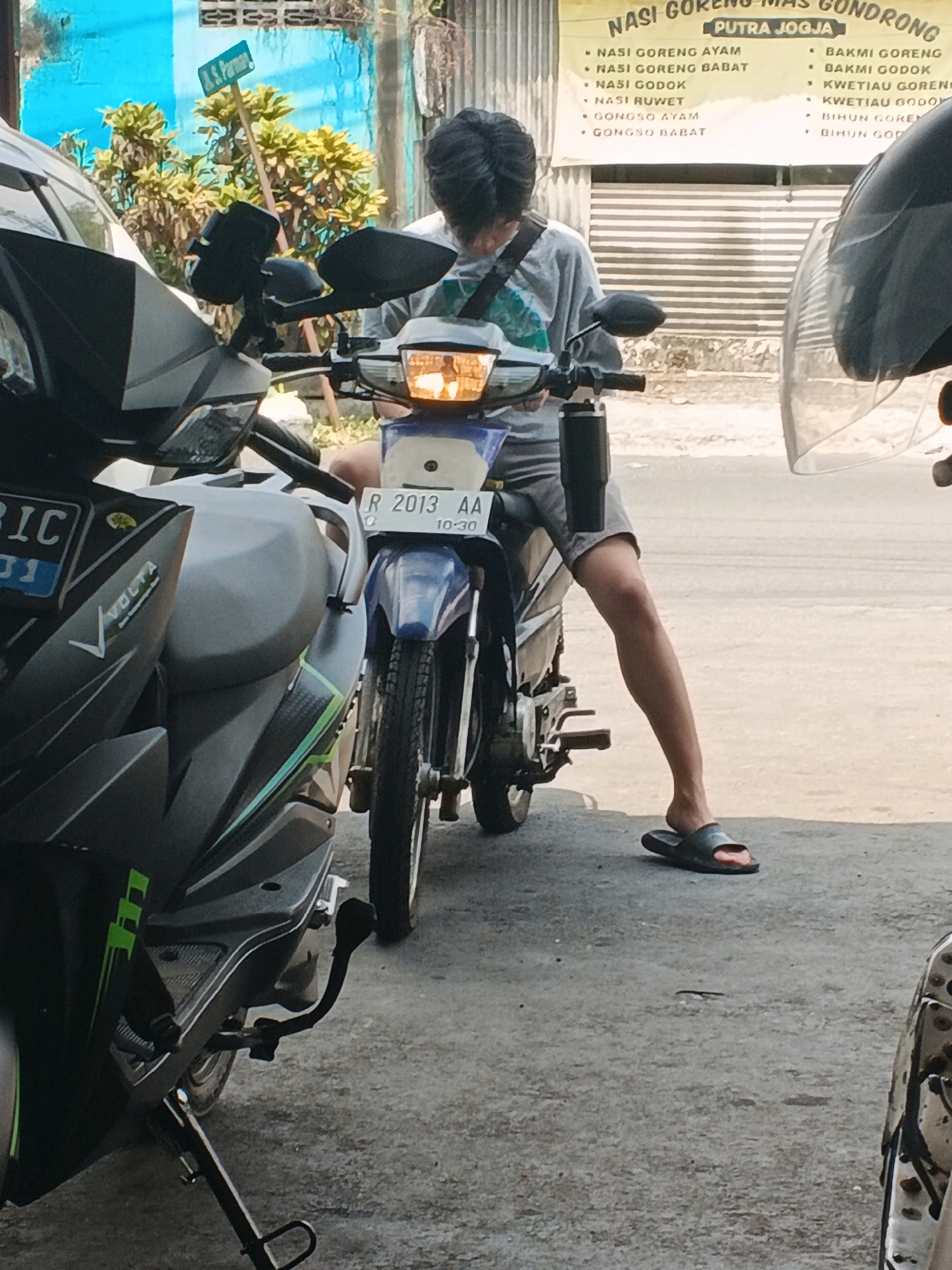 A young person sits on a motorcycle, facing right. They wear a gray t-shirt, gray shorts, and dark slides. The motorcycle is blue and white with a license plate reading "R 2013 AA". To the left, another motorcycle is partially visible, a dark gray scooter with green accents and a license plate reading "BIC 01". The background shows a yellow sign with text in Indonesian, a blue wall, and foliage. The scene appears to be in Genuk Barat, Indonesia.