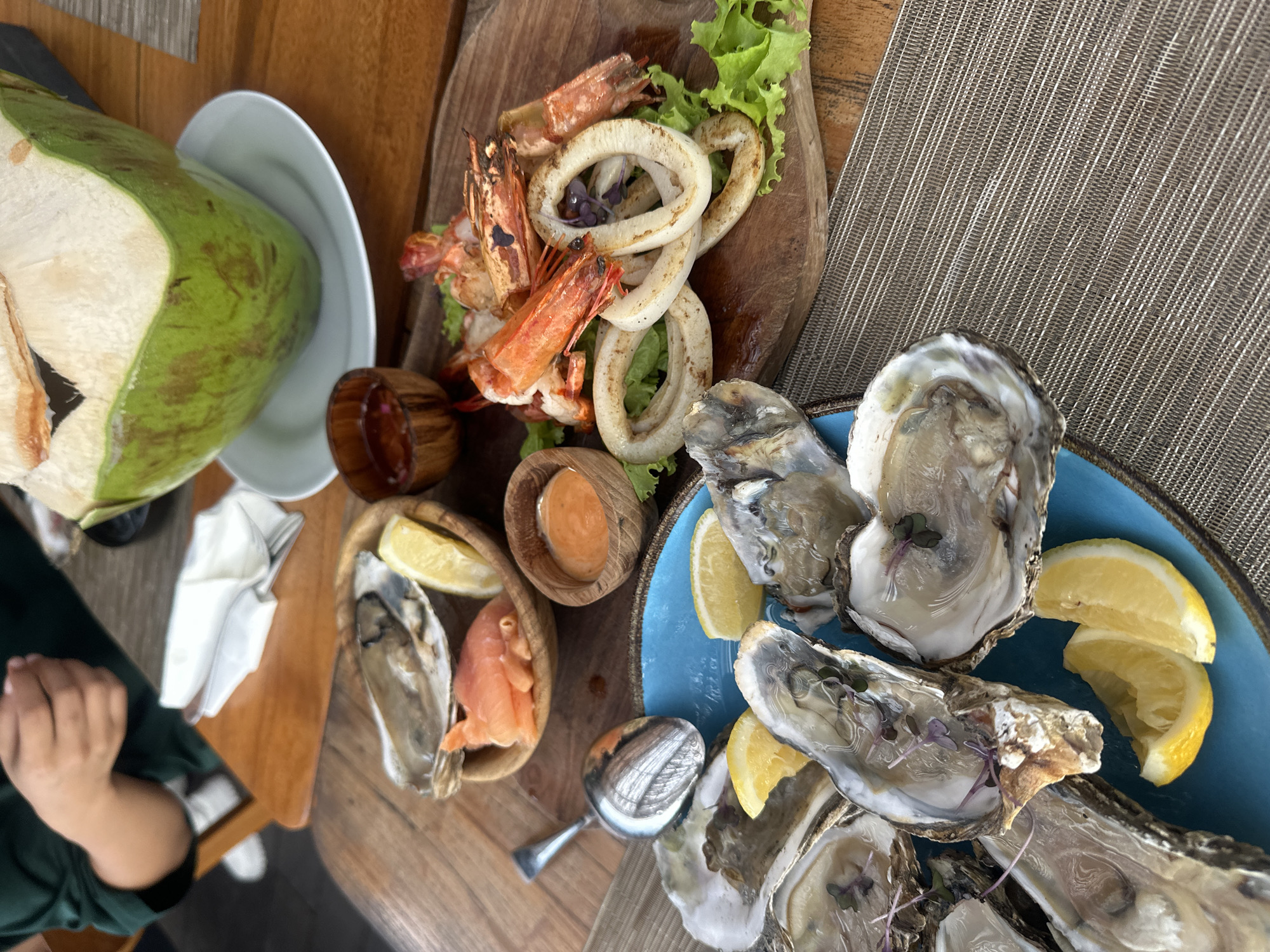 The image presents a seafood platter, likely served at a restaurant. On the left, a partially opened young coconut sits in a white bowl. Adjacent to it, a wooden board holds grilled shrimp and calamari rings, accompanied by lettuce and two small wooden bowls, one containing a sauce and the other a salmon slice and a lemon wedge. A blue plate holds multiple open oysters garnished with lemon wedges and small purple sprouts. The seafood rests on a textured, possibly woven, placemat. In the bottom left, part of a person's arm and hand are visible. The setting appears to be in North Kuta, Indonesia, as the meal combination of seafood and coconut is common in this region. The environment suggests outdoor dining, potentially in a tropical or coastal location.